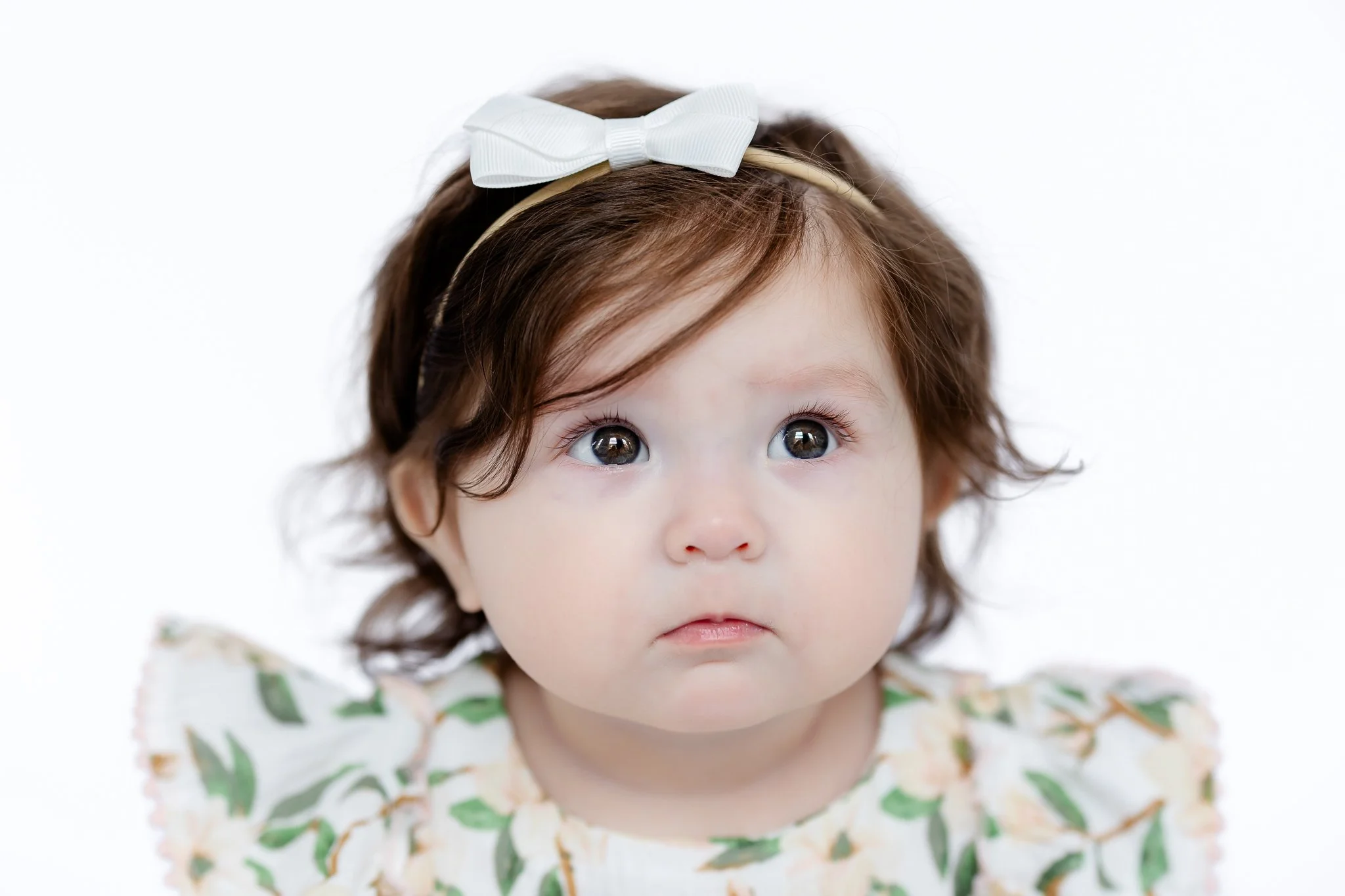 Baby with dark hair wearing a floral dress and a white bow headband, looking upwards.