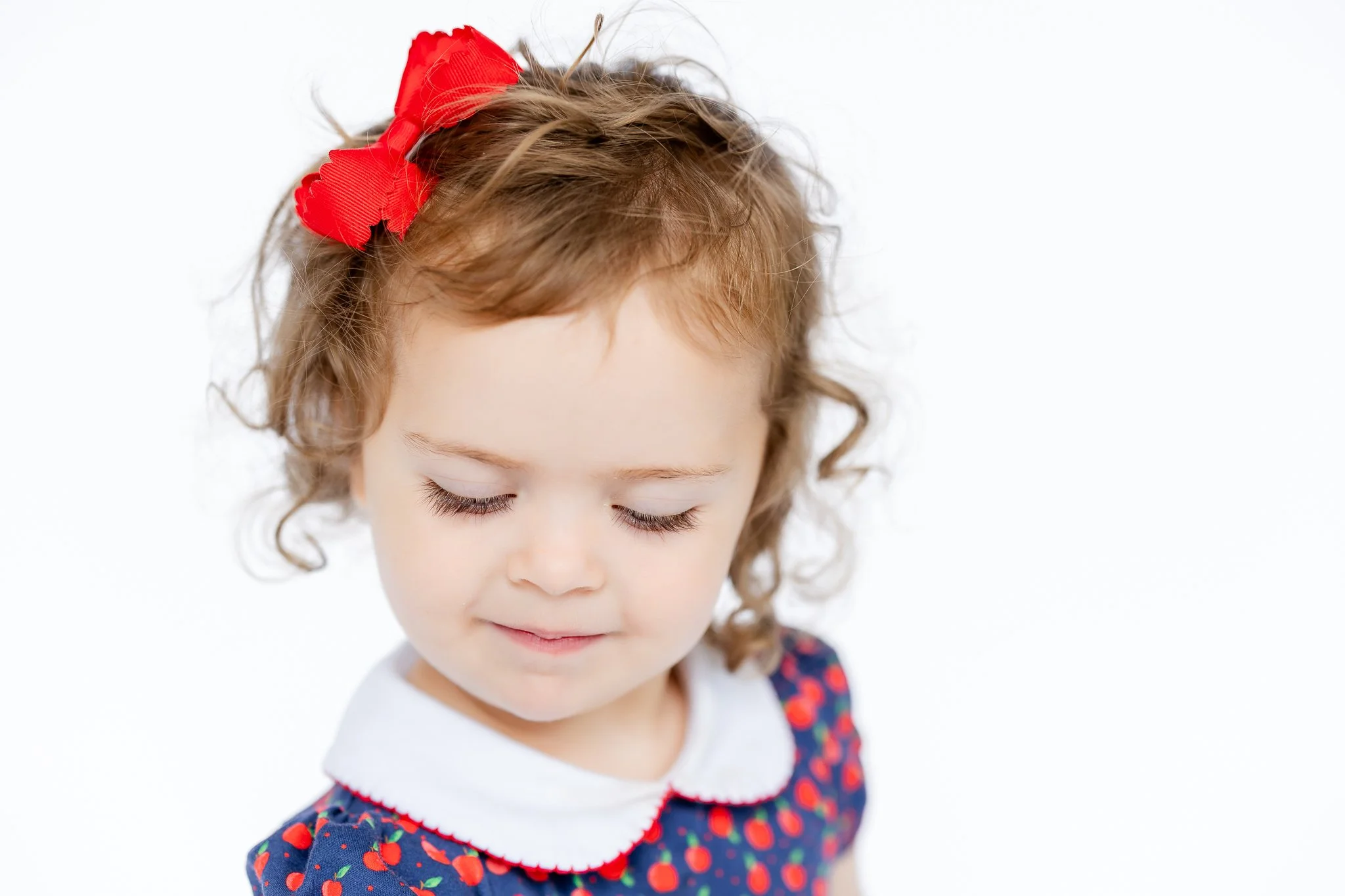 Young child with closed eyes, wearing a red hair bow and a dress with a fruit pattern.