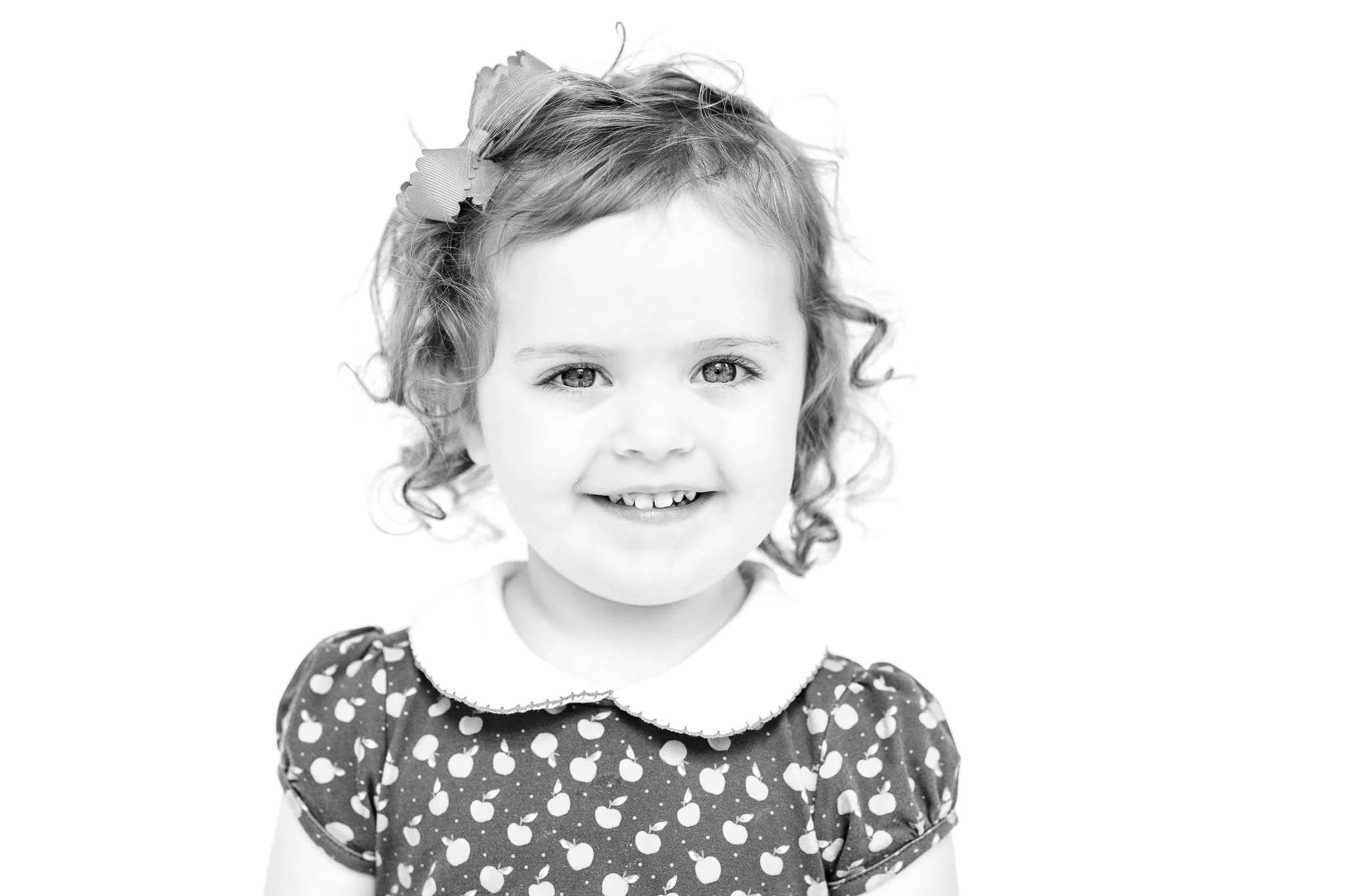 Black and white portrait of a young girl with curly hair and a bow, wearing a patterned dress with a collar, smiling at the camera.