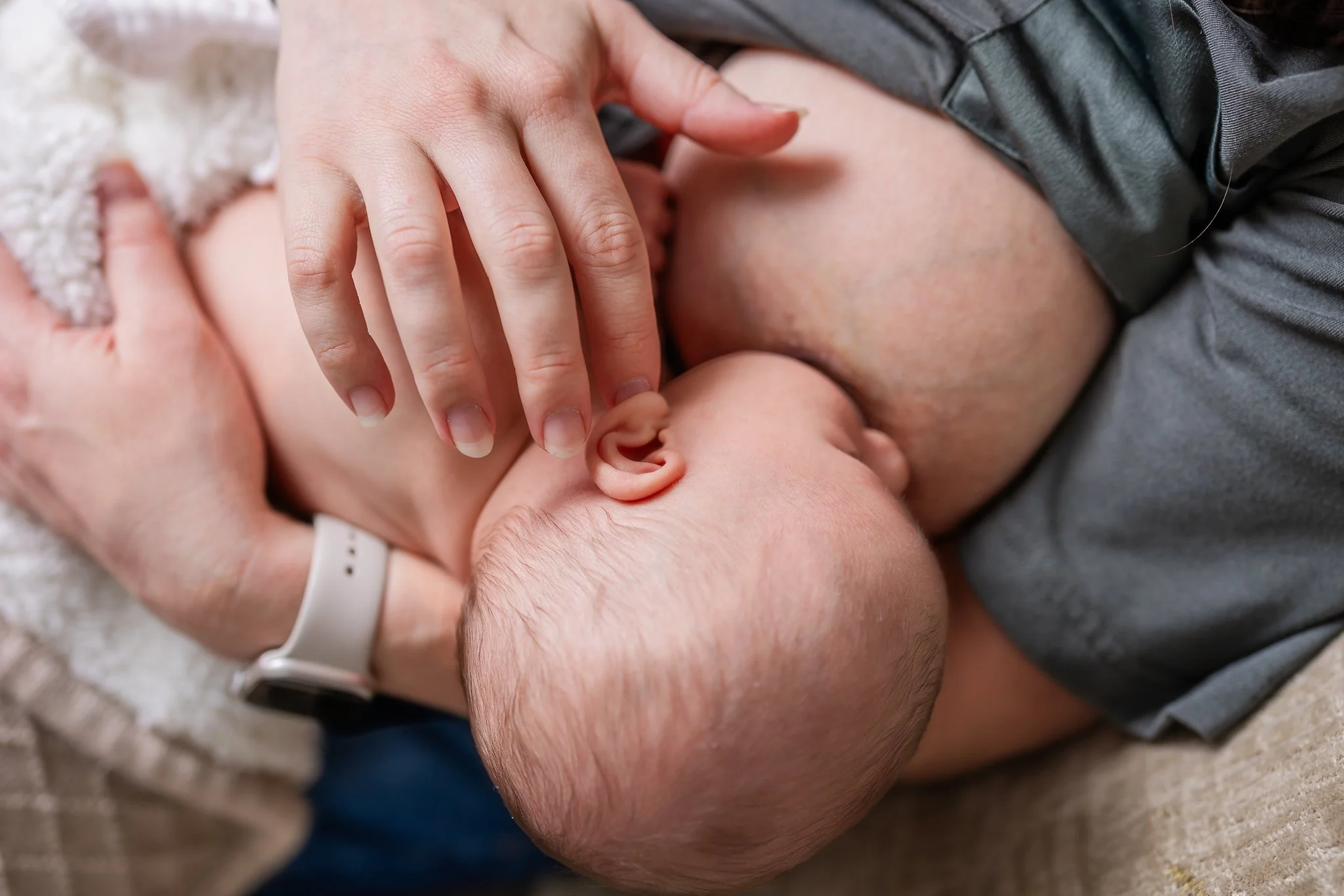 Close-up of a baby breastfeeding, with an adult's hand gently resting on the baby's head, wearing a watch.