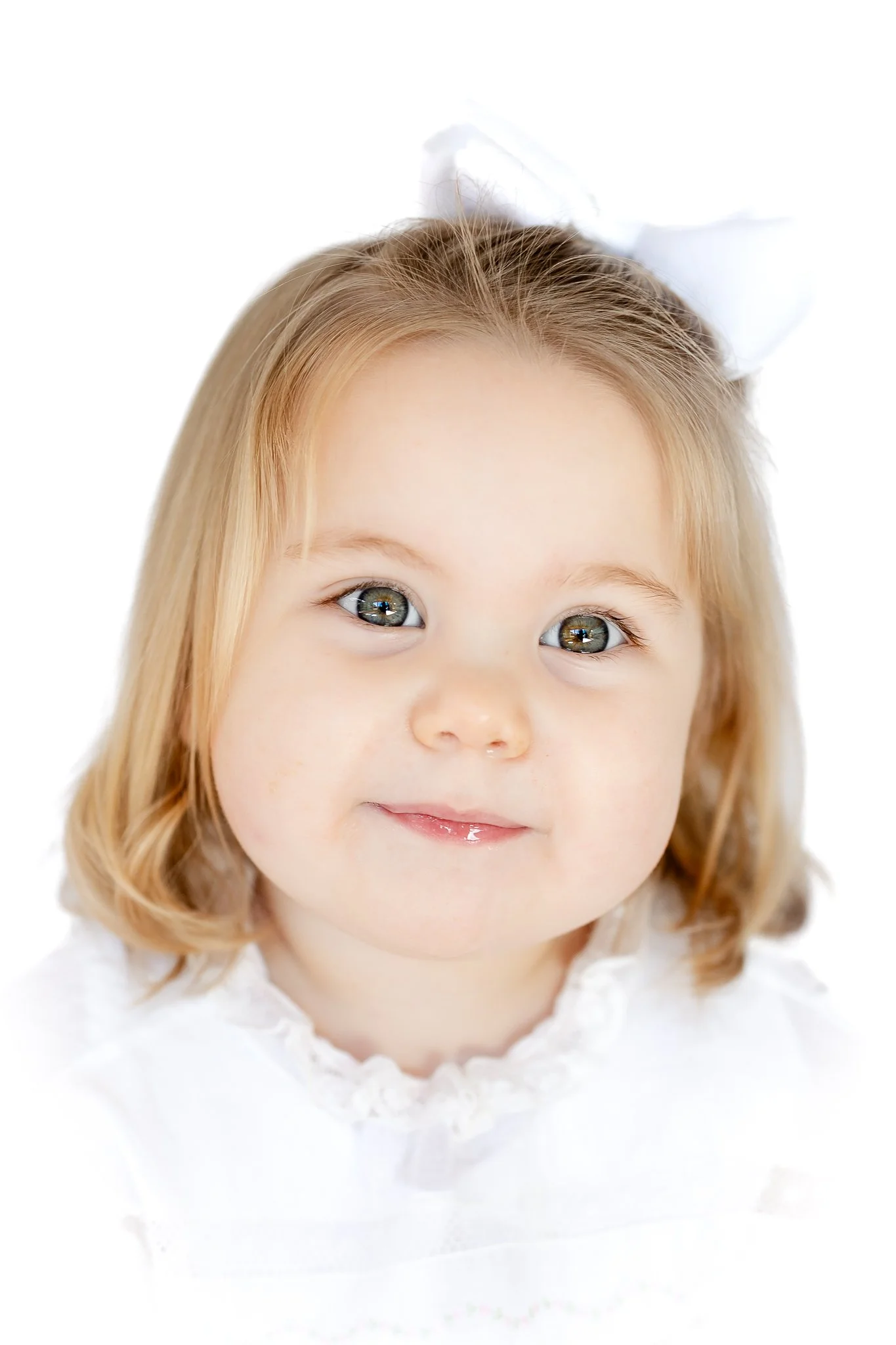 A young child with blonde hair and a white bow, wearing a white dress, smiling at the camera.