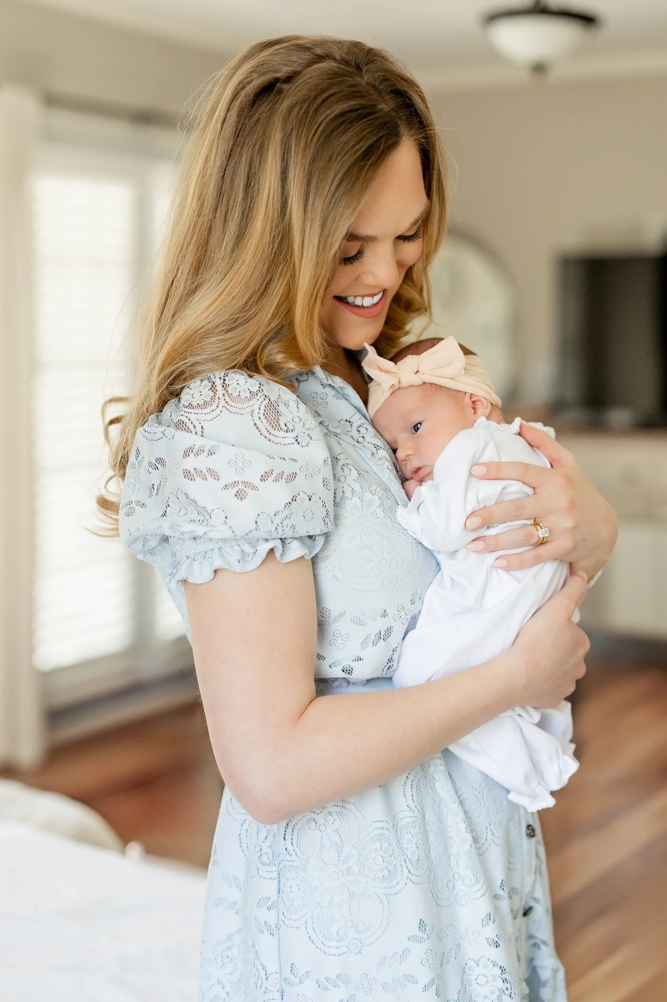 Woman smiling and holding a baby dressed in white with a headband in a softly lit room.