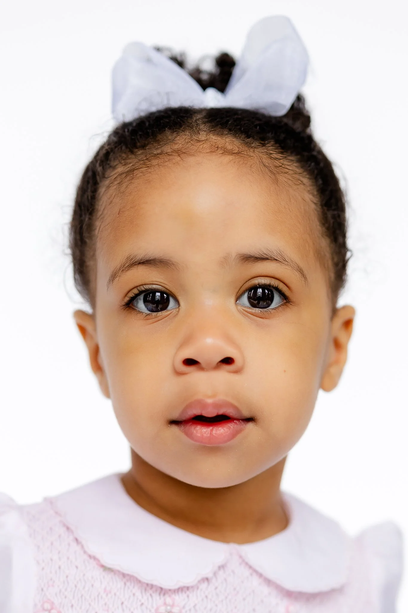 Portrait of a young child with a bow in their hair, wearing a light-colored outfit, against a white background.