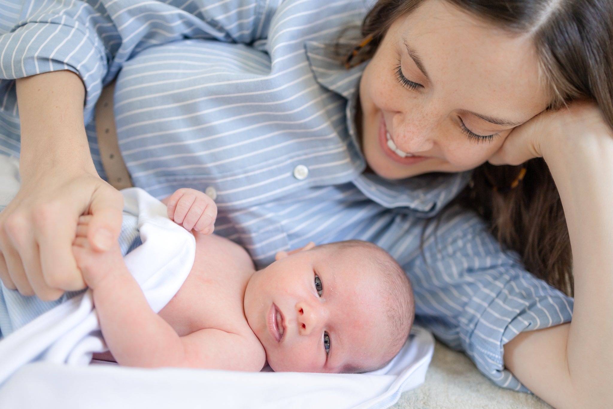 A smiling woman lying next to a newborn baby wrapped in a white blanket, holding the baby's hand.