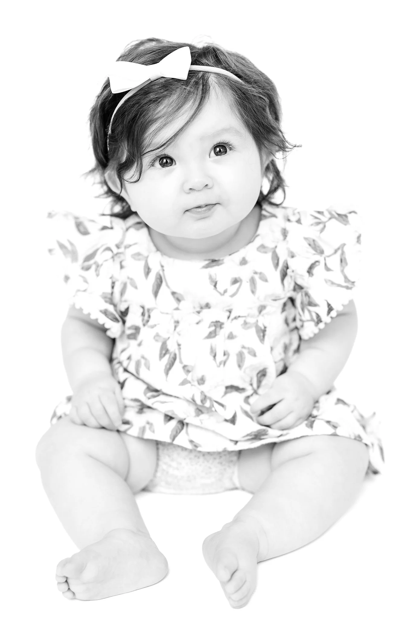 Black and white photo of a baby with wavy hair and a bow headband, wearing a floral dress, sitting on the floor.