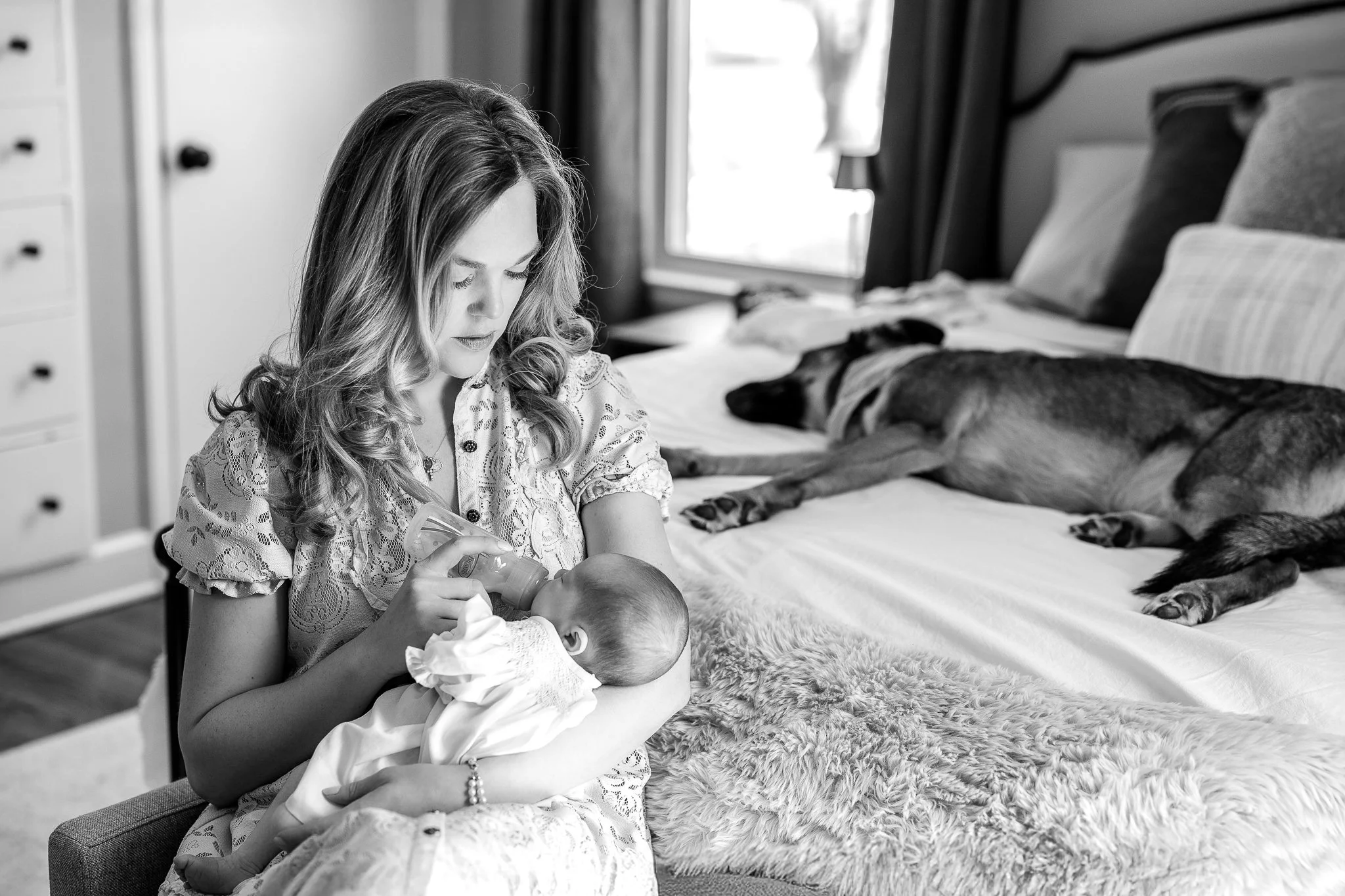 Woman bottle-feeding a baby while sitting on a chair beside a bed where a dog is lying down.