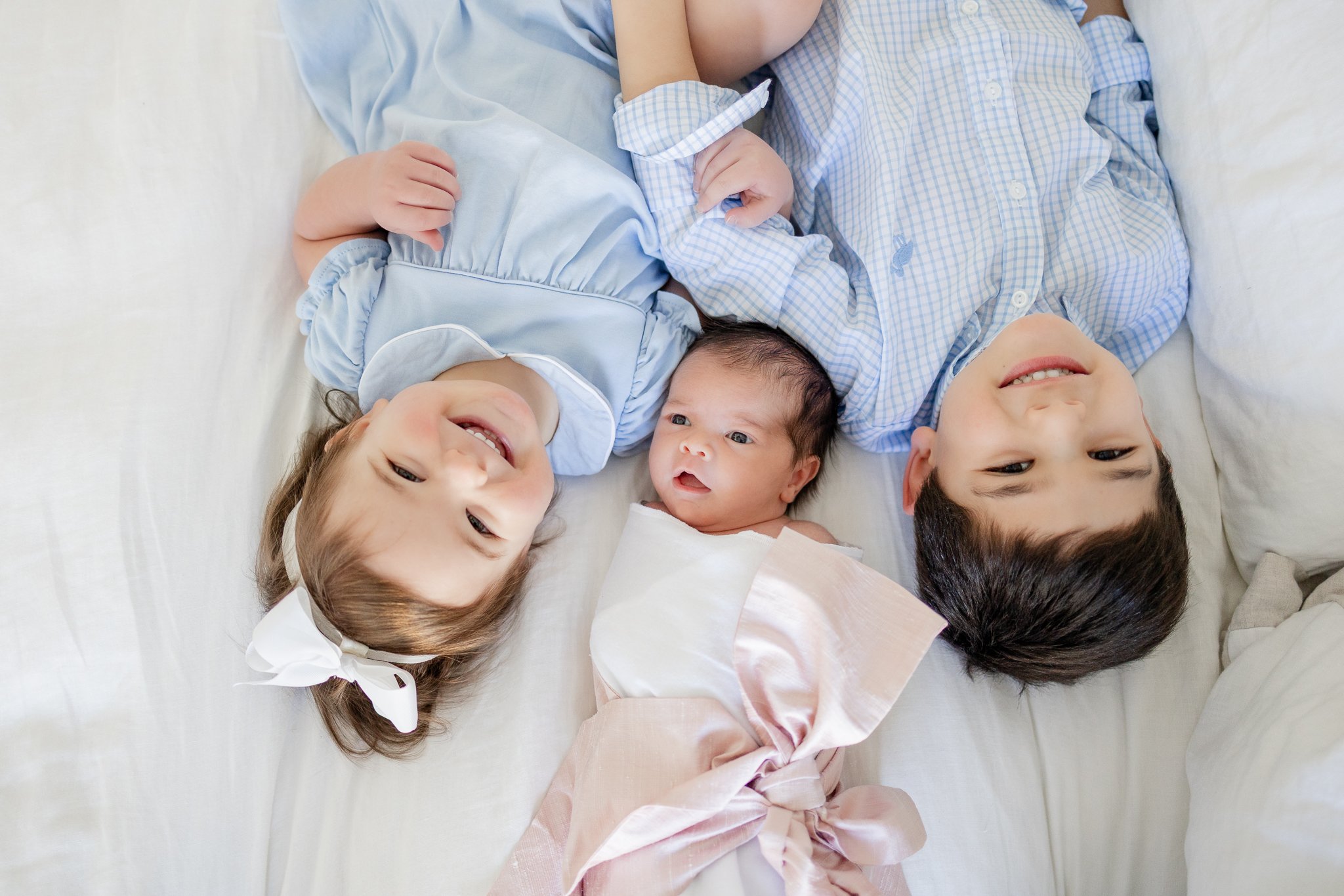 Three children lying on a bed, including a newborn wrapped in a pink blanket with two older siblings, a boy and a girl, smiling and dressed in light blue.