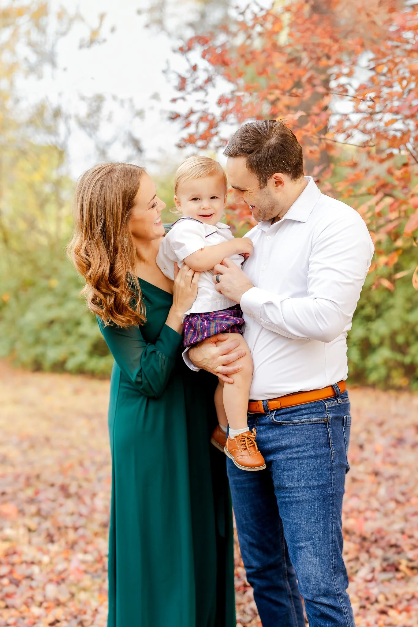 A family with a mother, father, and child standing outdoors in a park during fall. The mother is wearing a green dress, the father is in a white shirt and jeans, and they are holding a young child wearing a white shirt and plaid shorts. The backgroun