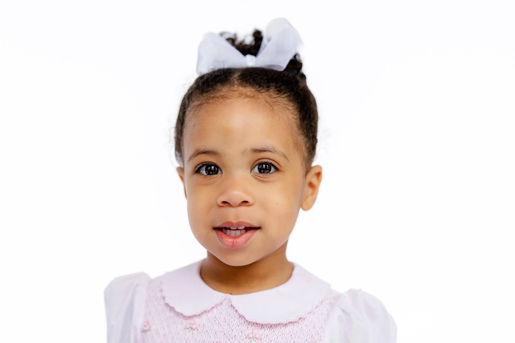 Toddler girl with a white ribbon in her hair, wearing a pink dress, smiling at the camera.