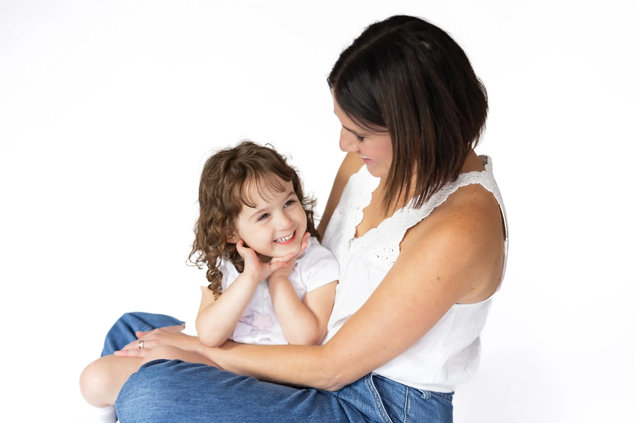A woman holding a smiling child on her lap, both wearing white tops, sitting against a white background.