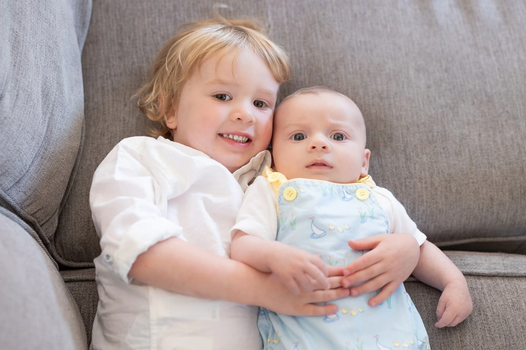 Two children lying on a gray couch; a toddler hugging a baby, both wearing light-colored clothes.