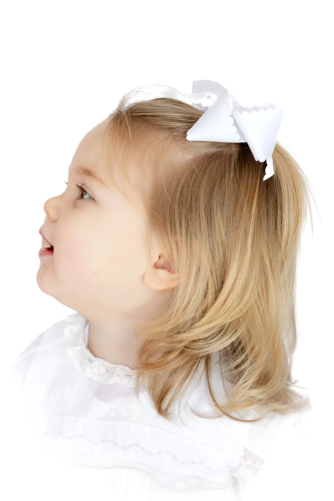 Profile of a young child with blonde hair and a white bow, wearing a white outfit.
