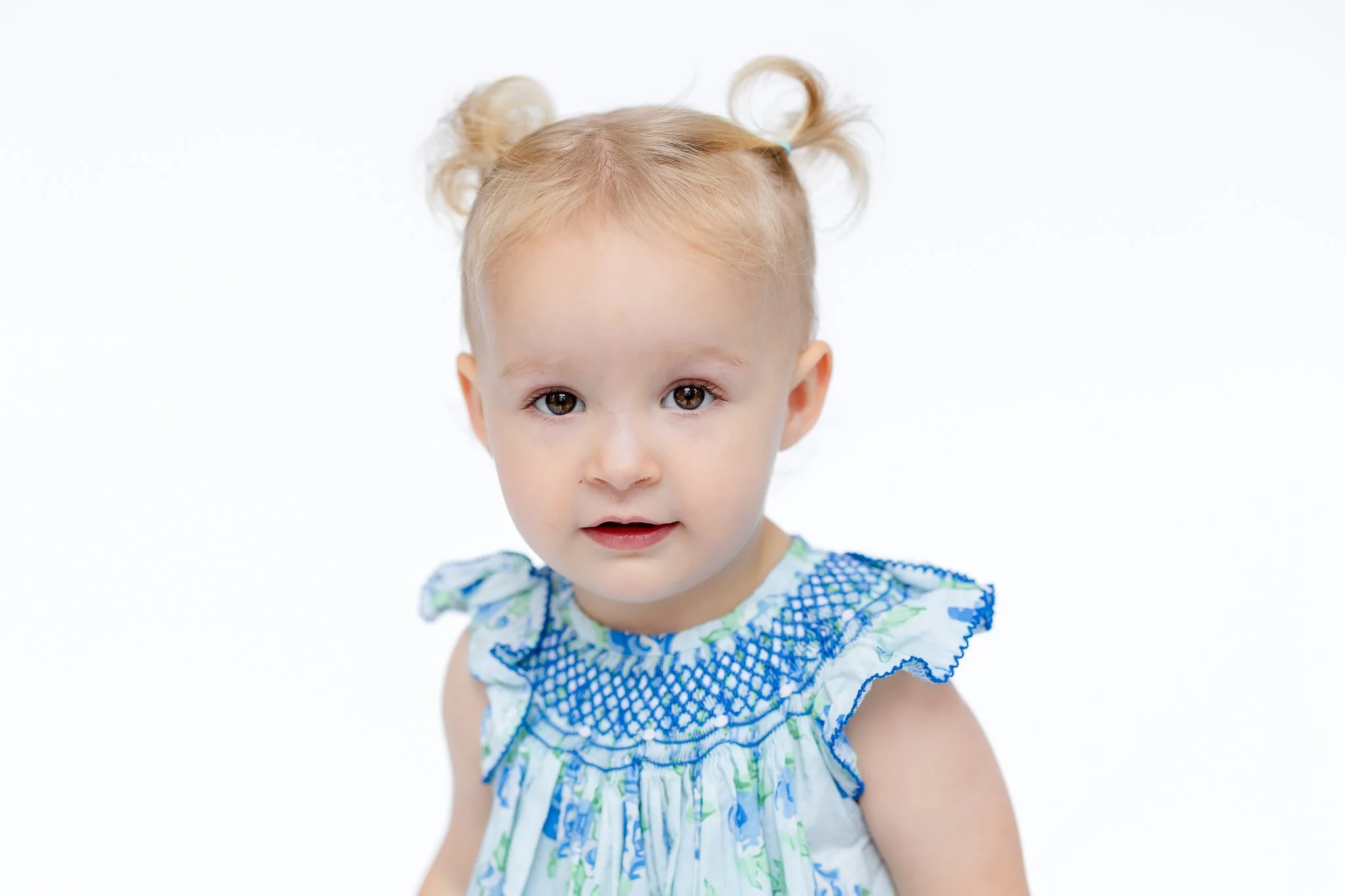 Toddler with light hair styled in two small buns, wearing a blue and white dress with a floral pattern on a white background.