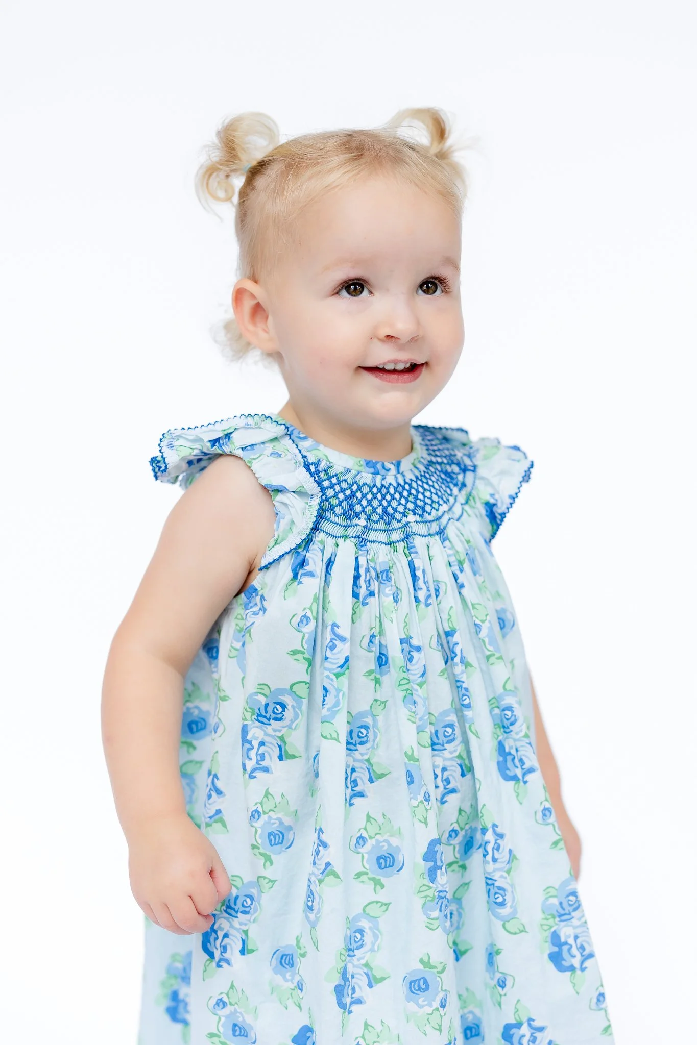 Smiling toddler in a blue floral dress with ruffled sleeves, wearing her hair in two small buns.