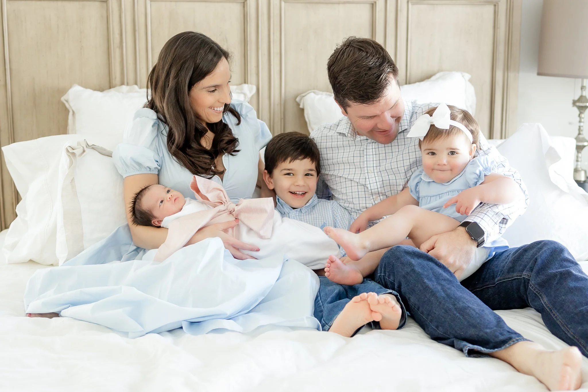 Family of five sitting on a bed, with two parents, a young child, a toddler, and a newborn baby, all smiling and dressed in light blue and white clothing.