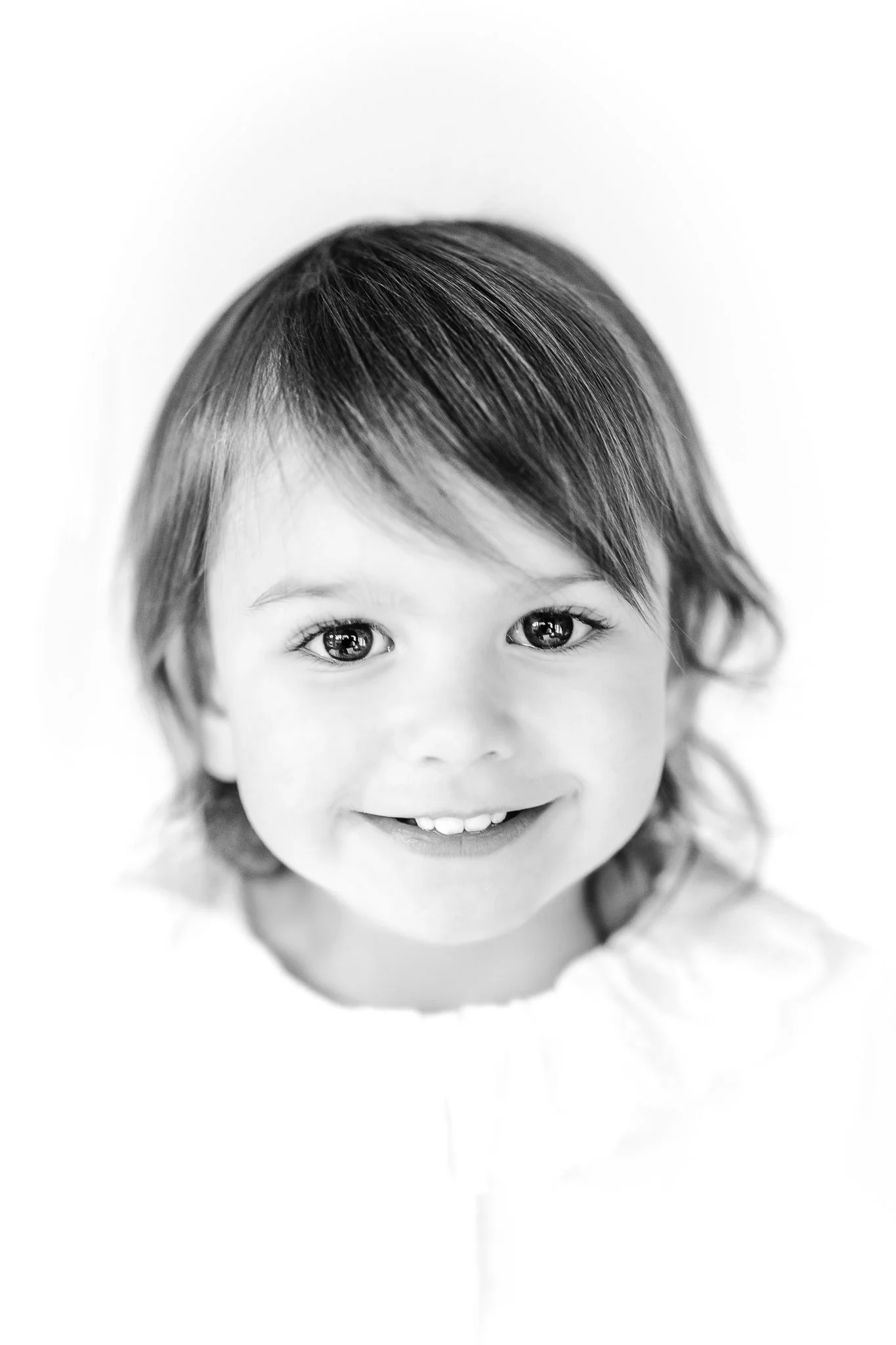 Black and white portrait of a smiling young child with medium-length hair and bright eyes, wearing a light-colored top.