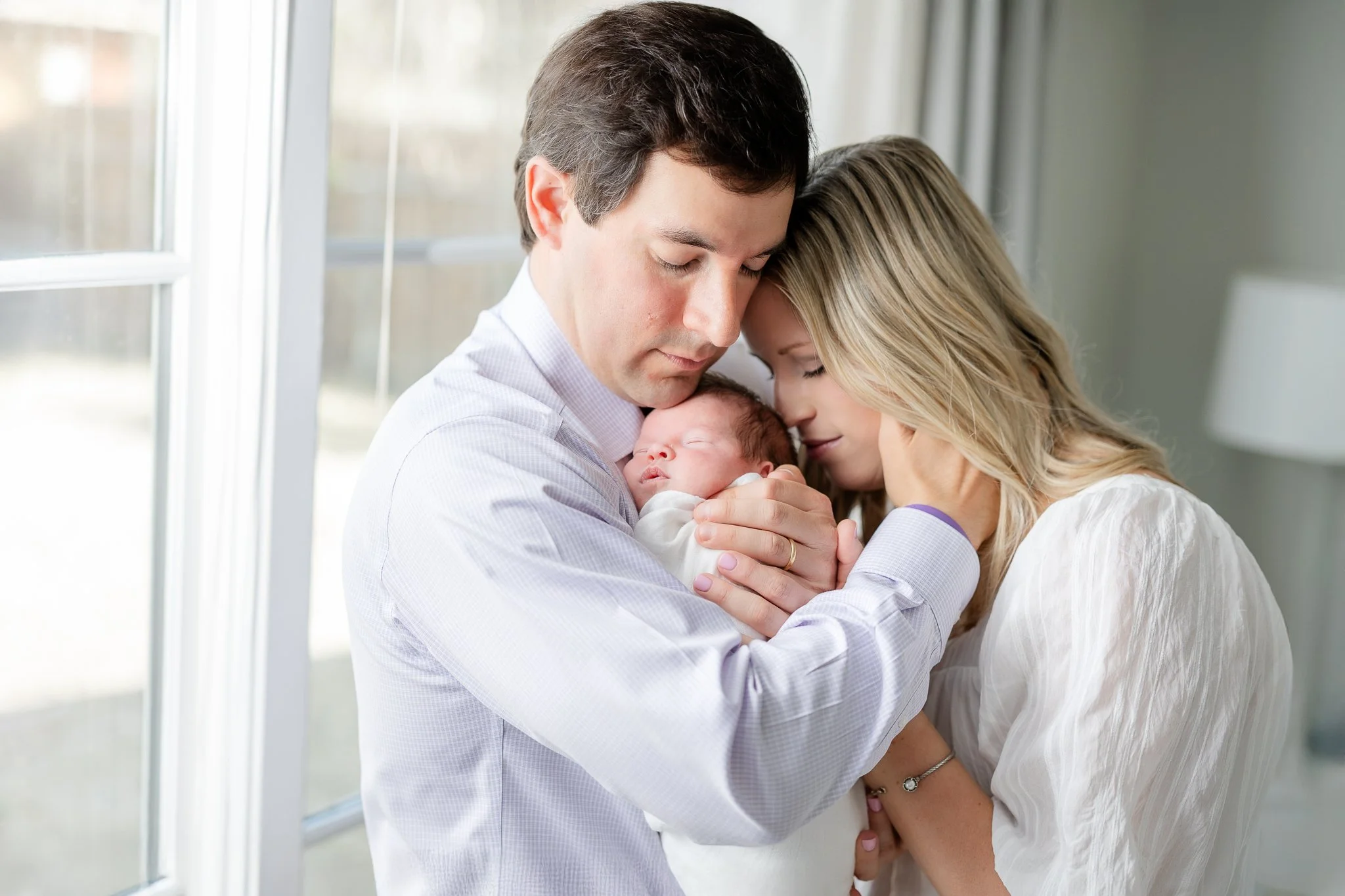 A loving couple holding a sleeping newborn baby in a warm embrace by a window.