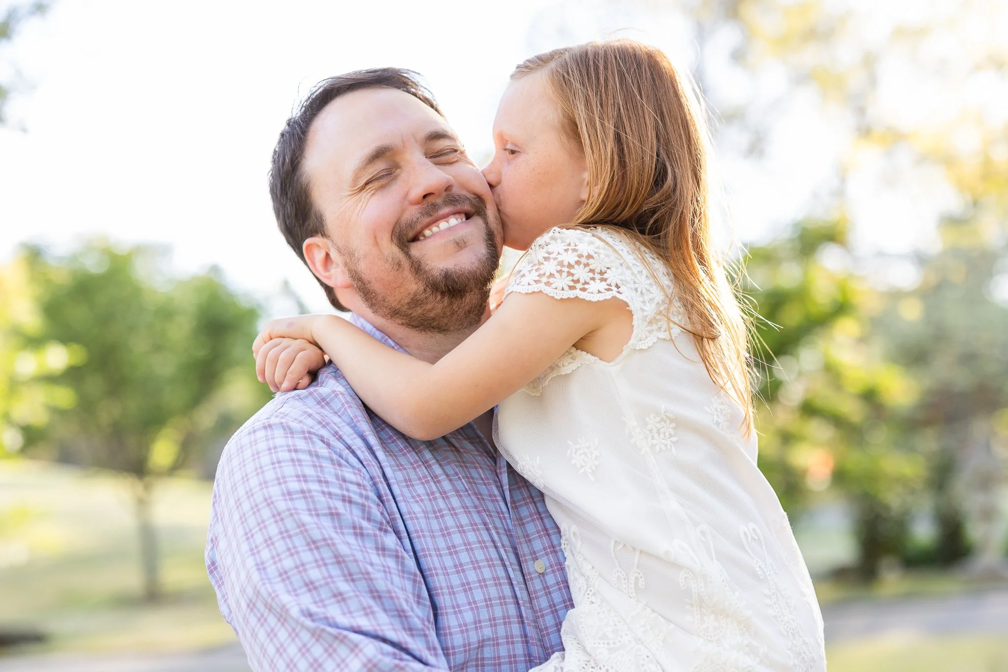 A smiling man with a beard, wearing a plaid shirt, holding a young girl in a white dress who is kissing his cheek. They are outdoors with blurred trees in the background.