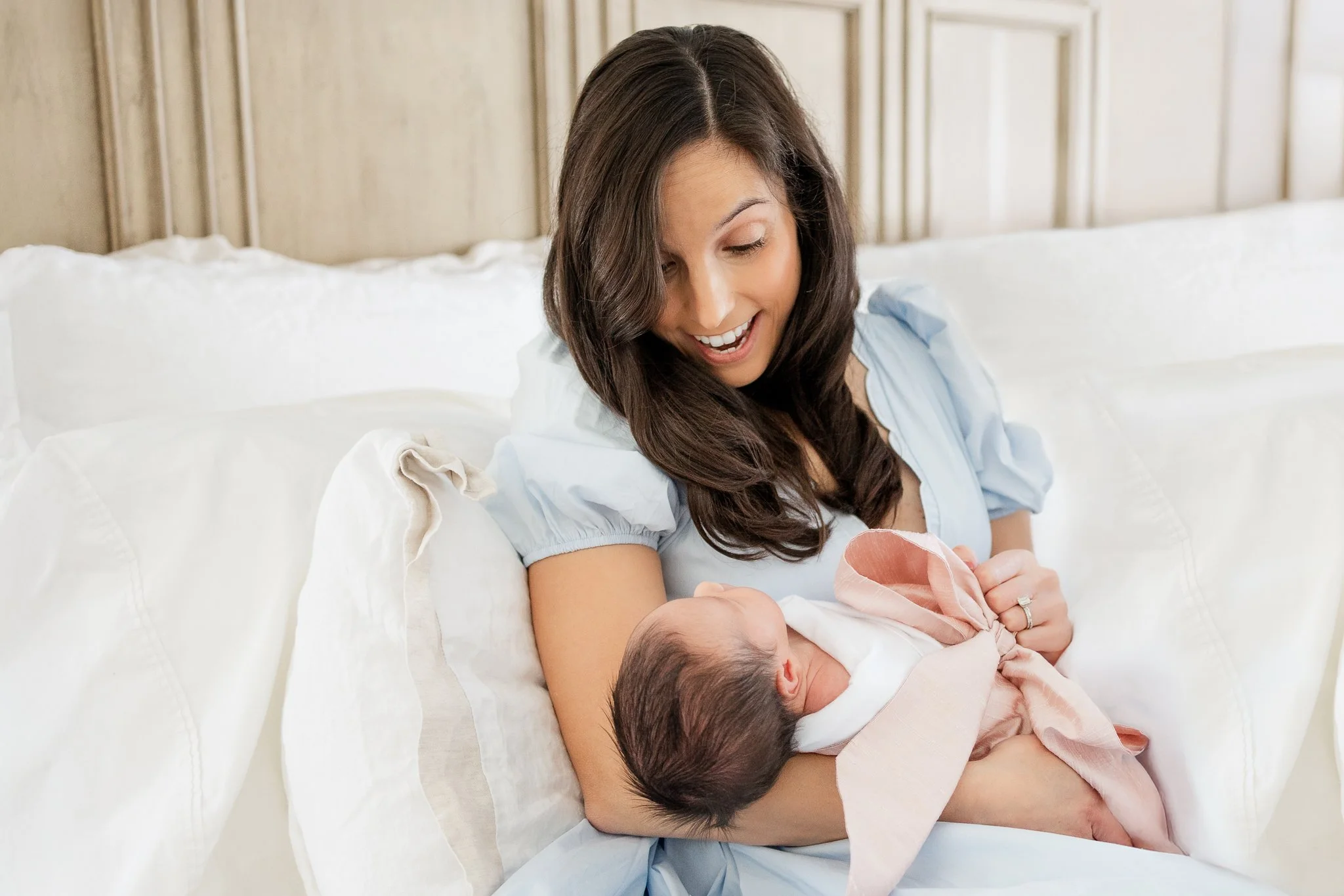 Woman holding a newborn wrapped in a pink blanket, sitting on a bed with white pillows.