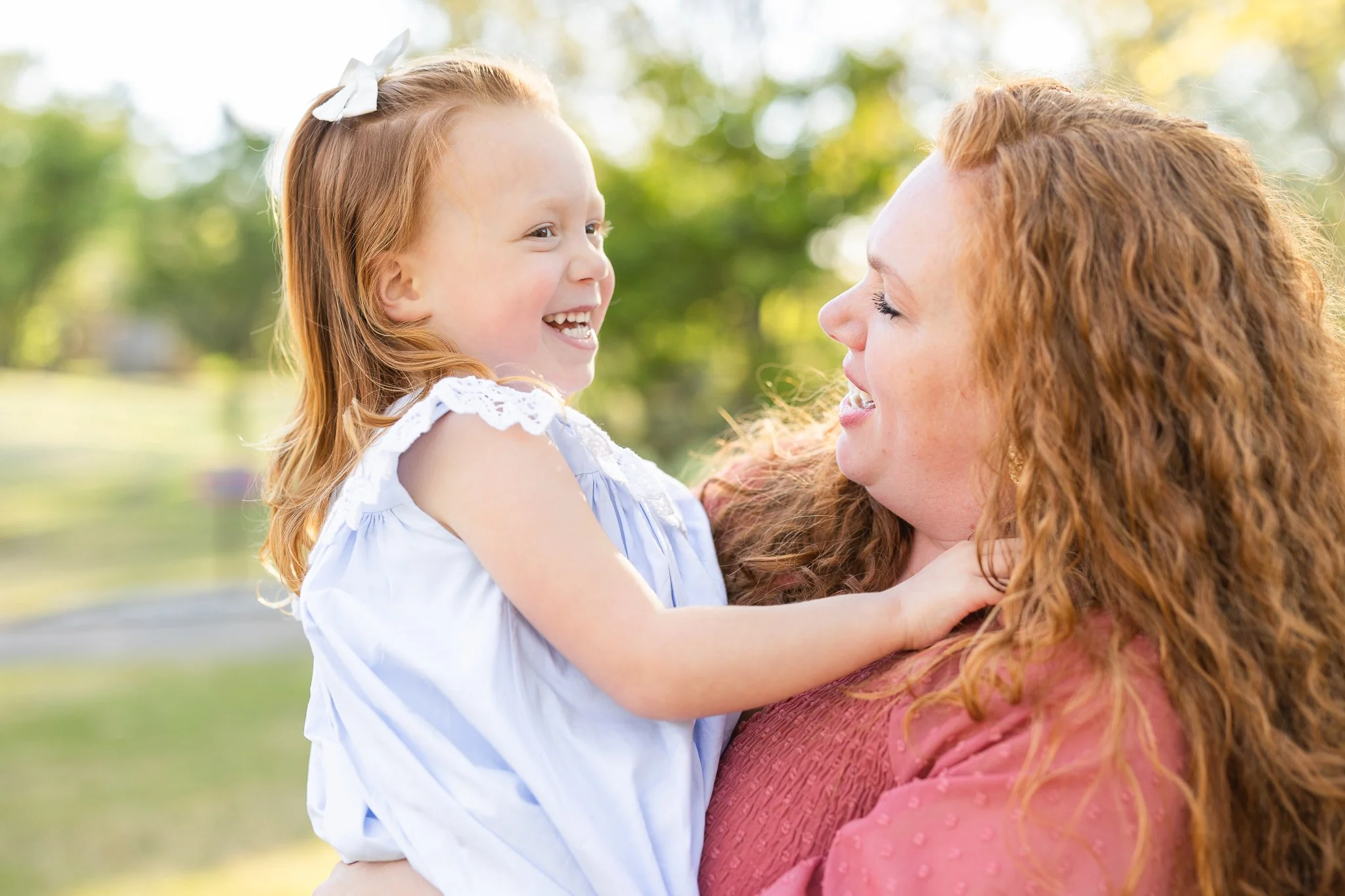 A woman holding a smiling young girl outdoors, both with red hair. The woman wears a red top, and the girl wears a light blue dress. They are laughing and enjoying the sunny day in a green park.