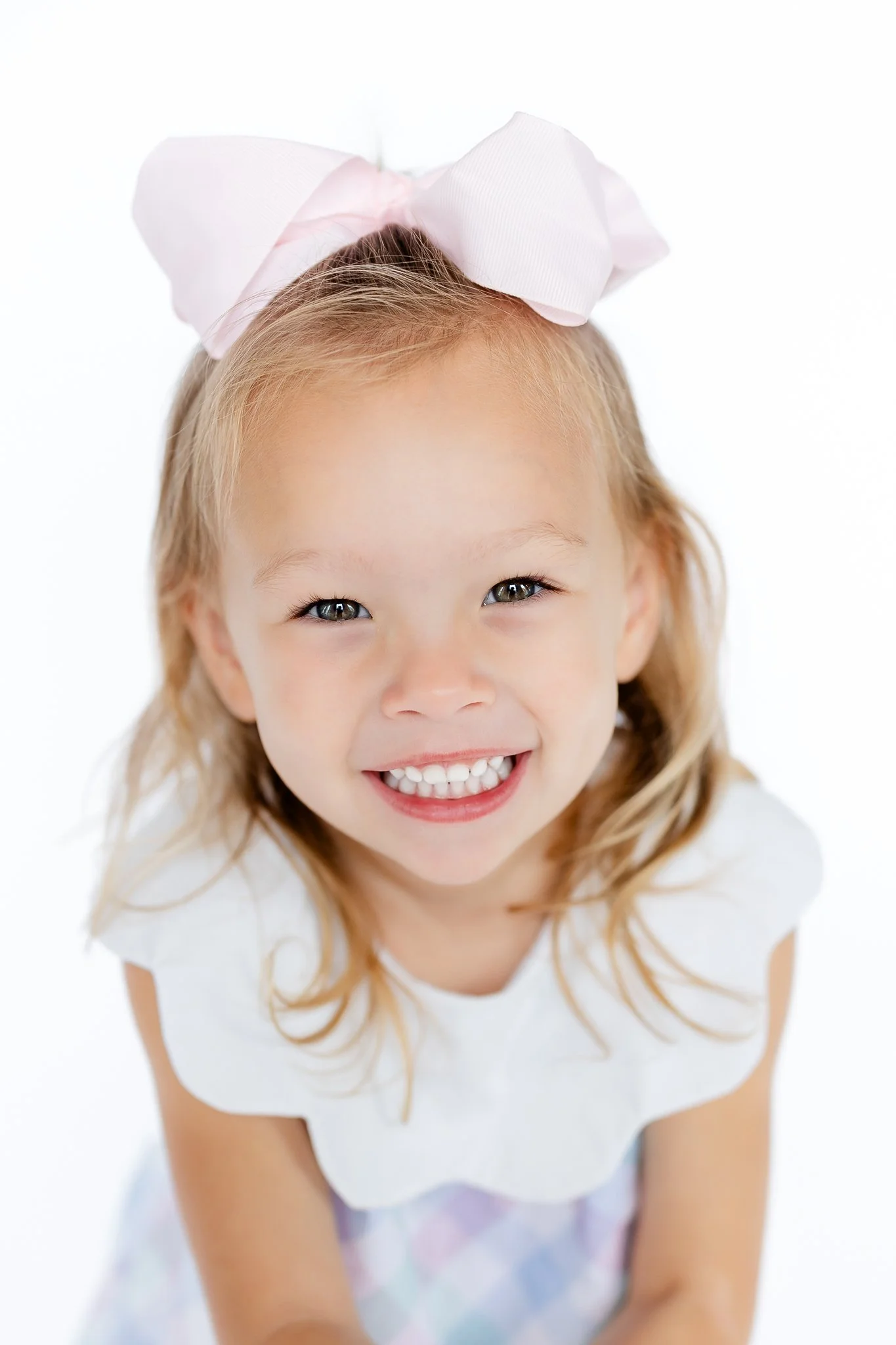 Smiling young child with a large bow in her hair, wearing a white dress with a pastel checkered pattern.