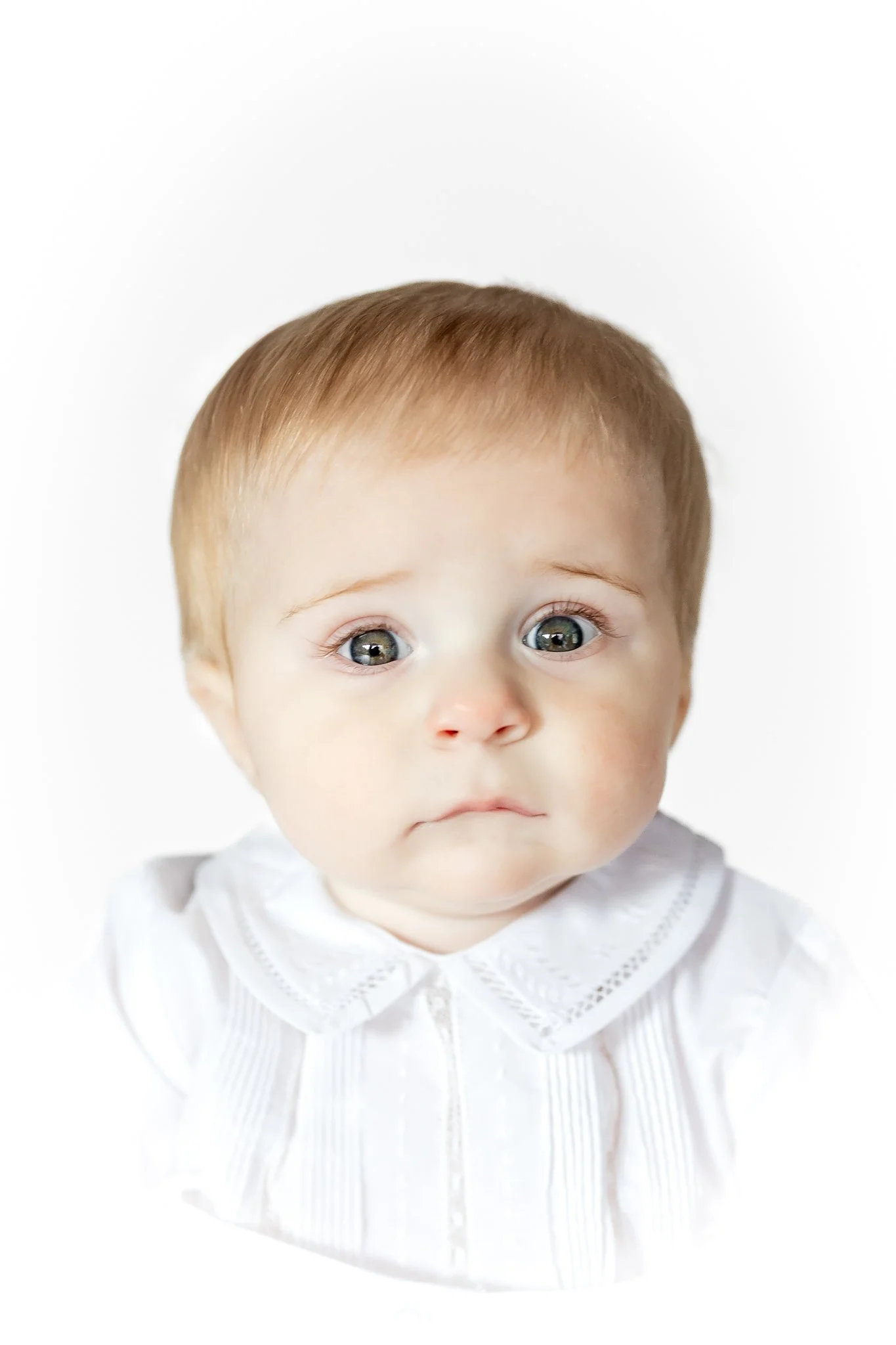 Portrait of a baby with a white shirt and neutral expression, with light brown hair and wide eyes.