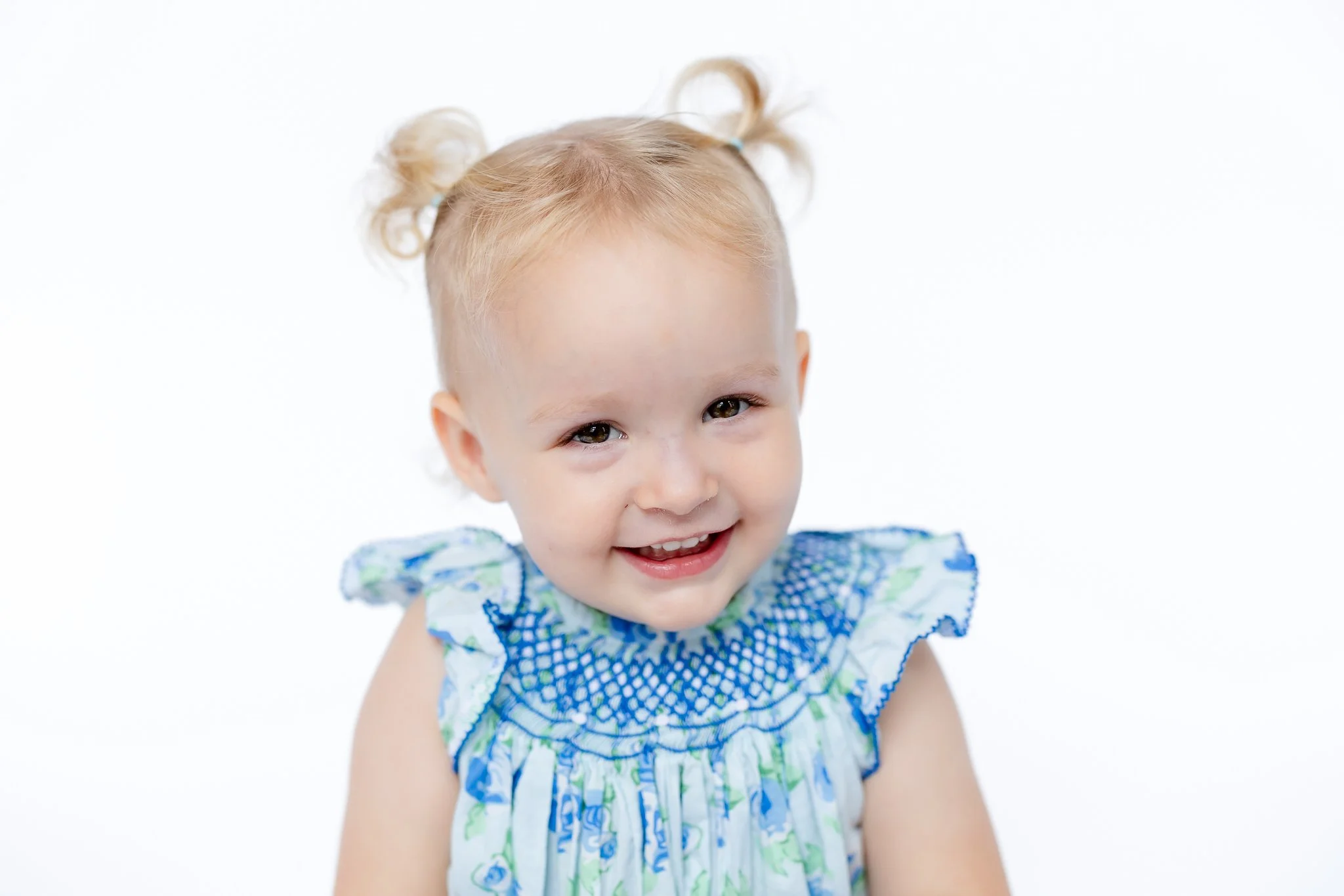 Smiling toddler with pigtails wearing a blue and white floral dress on a white background.