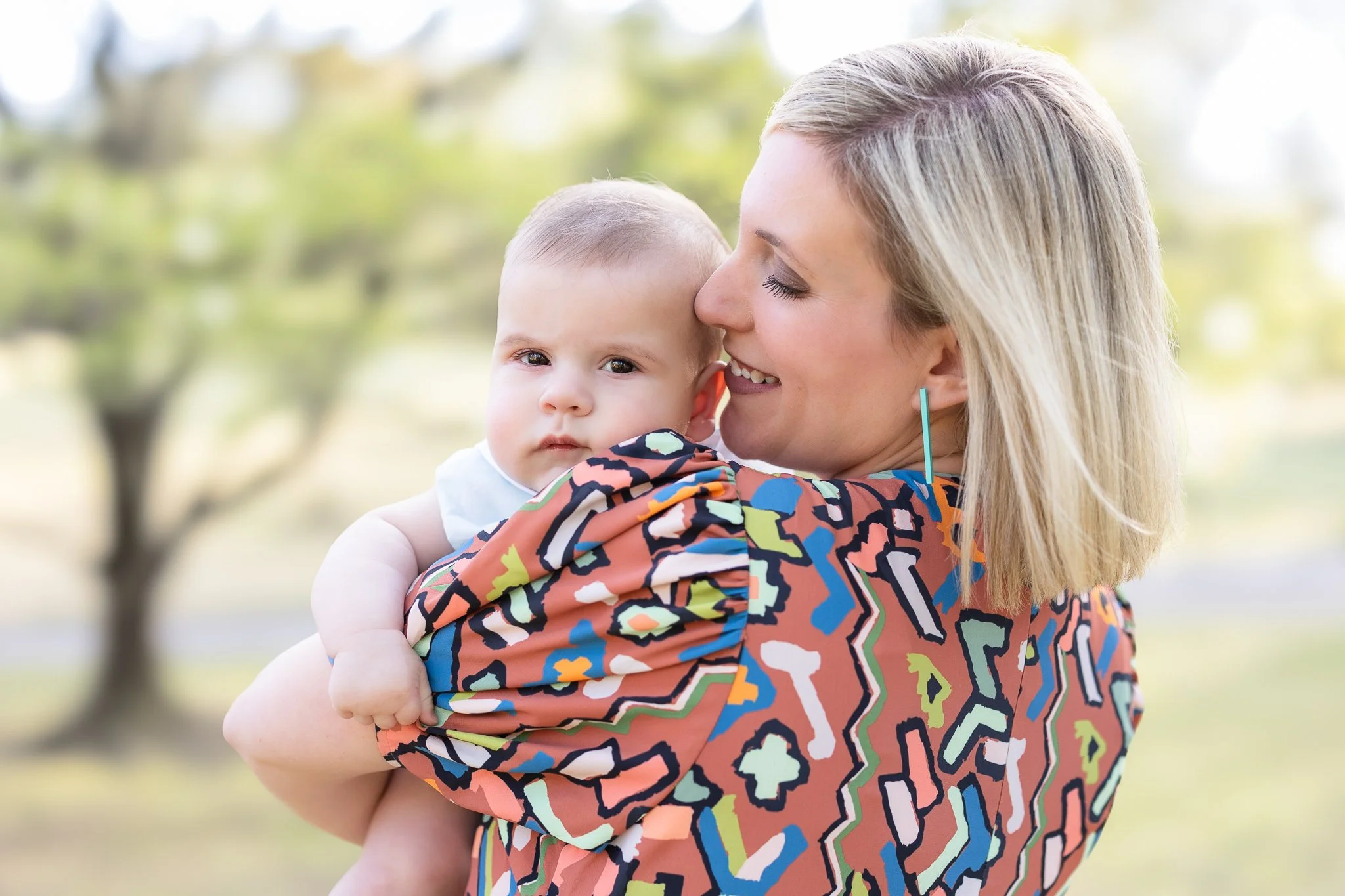 A woman holding a baby outdoors, with the woman's face turned towards the baby, both looking content. She wears a colorful patterned shirt and has short blonde hair. The background is blurred, suggesting a natural setting.