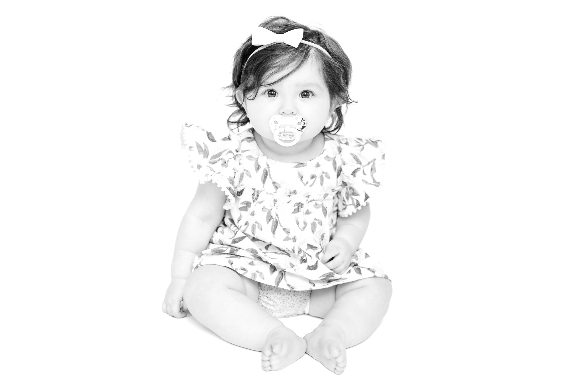 Black and white photo of a baby sitting, wearing a floral dress and a bow headband, with a pacifier.