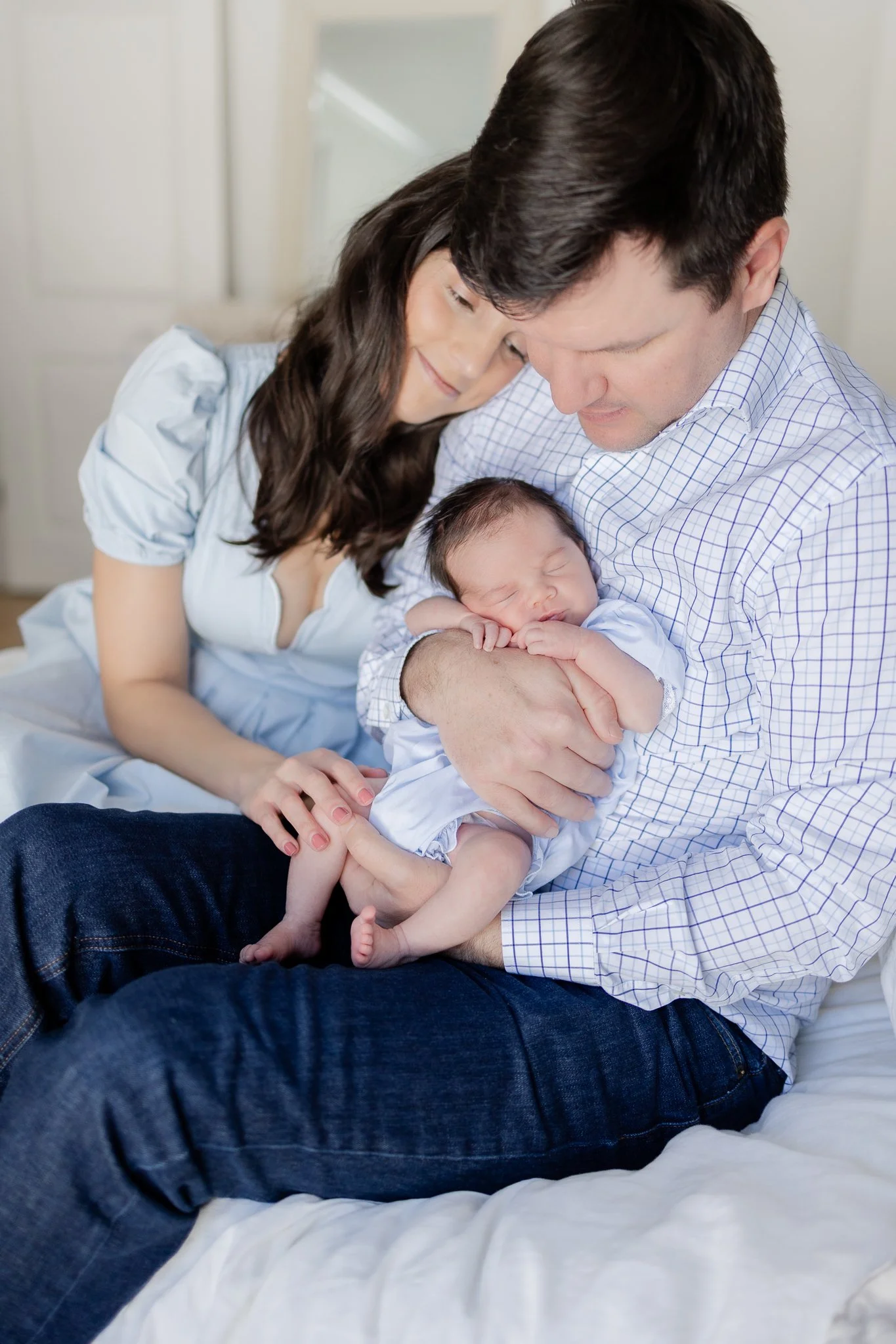 A couple sitting closely together on a bed, lovingly holding a sleeping newborn baby in their arms. The man is wearing a checkered shirt and jeans, while the woman wears a light blue dress.