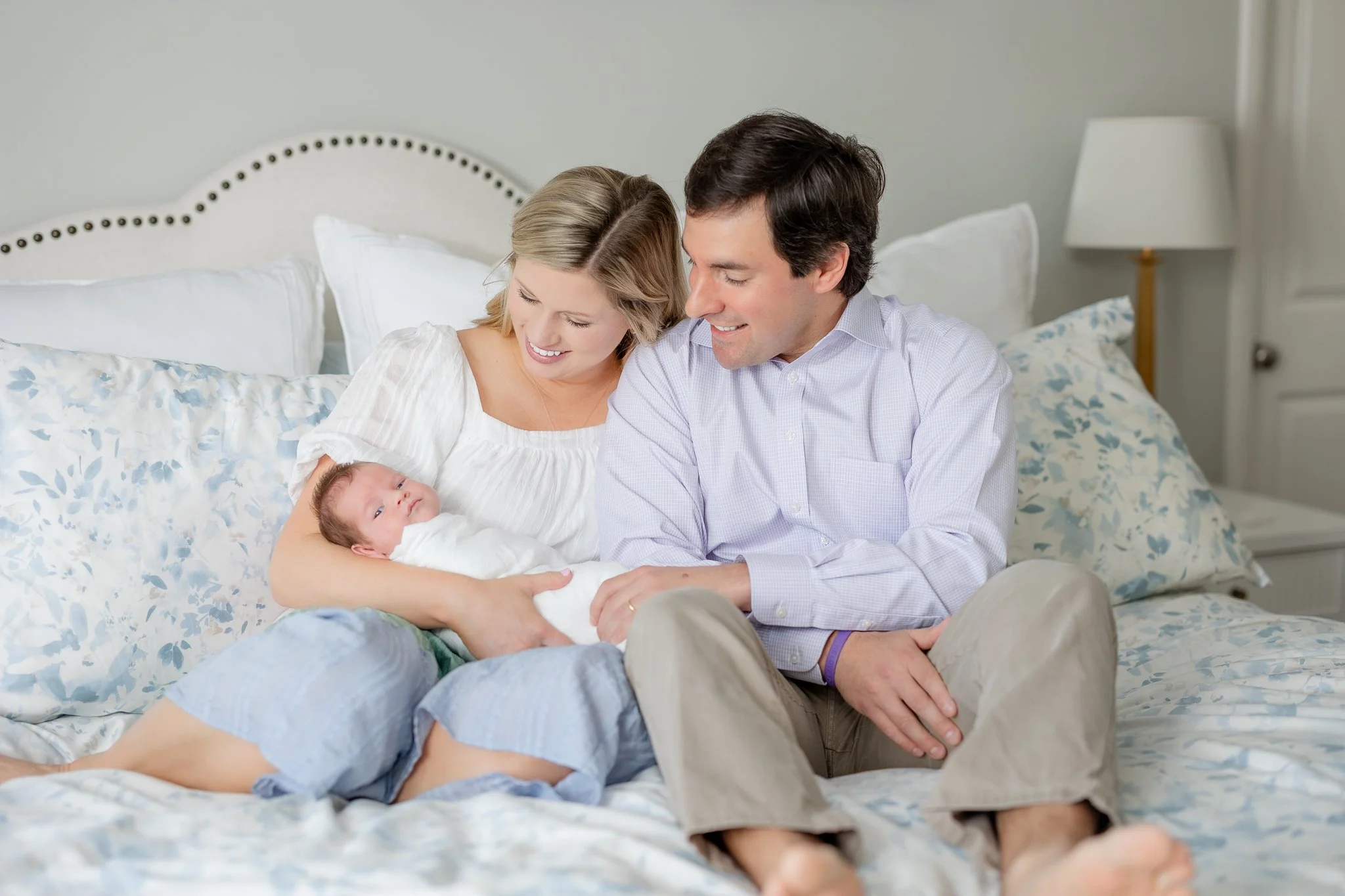 A couple sitting on a bed with a newborn baby between them. They are smiling and looking at the baby. The bed has floral-patterned bedding.
