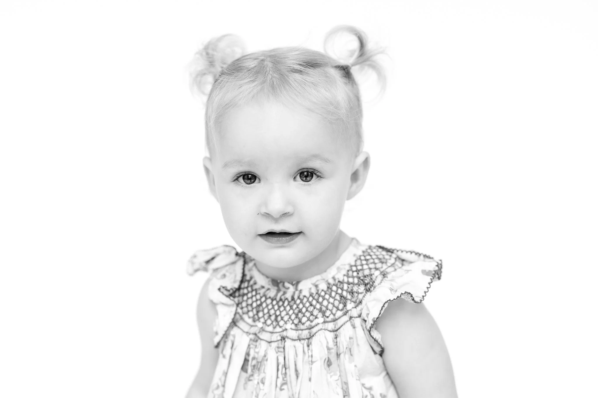 Black and white portrait of a toddler with pigtails wearing a patterned dress, looking at the camera on a plain background.