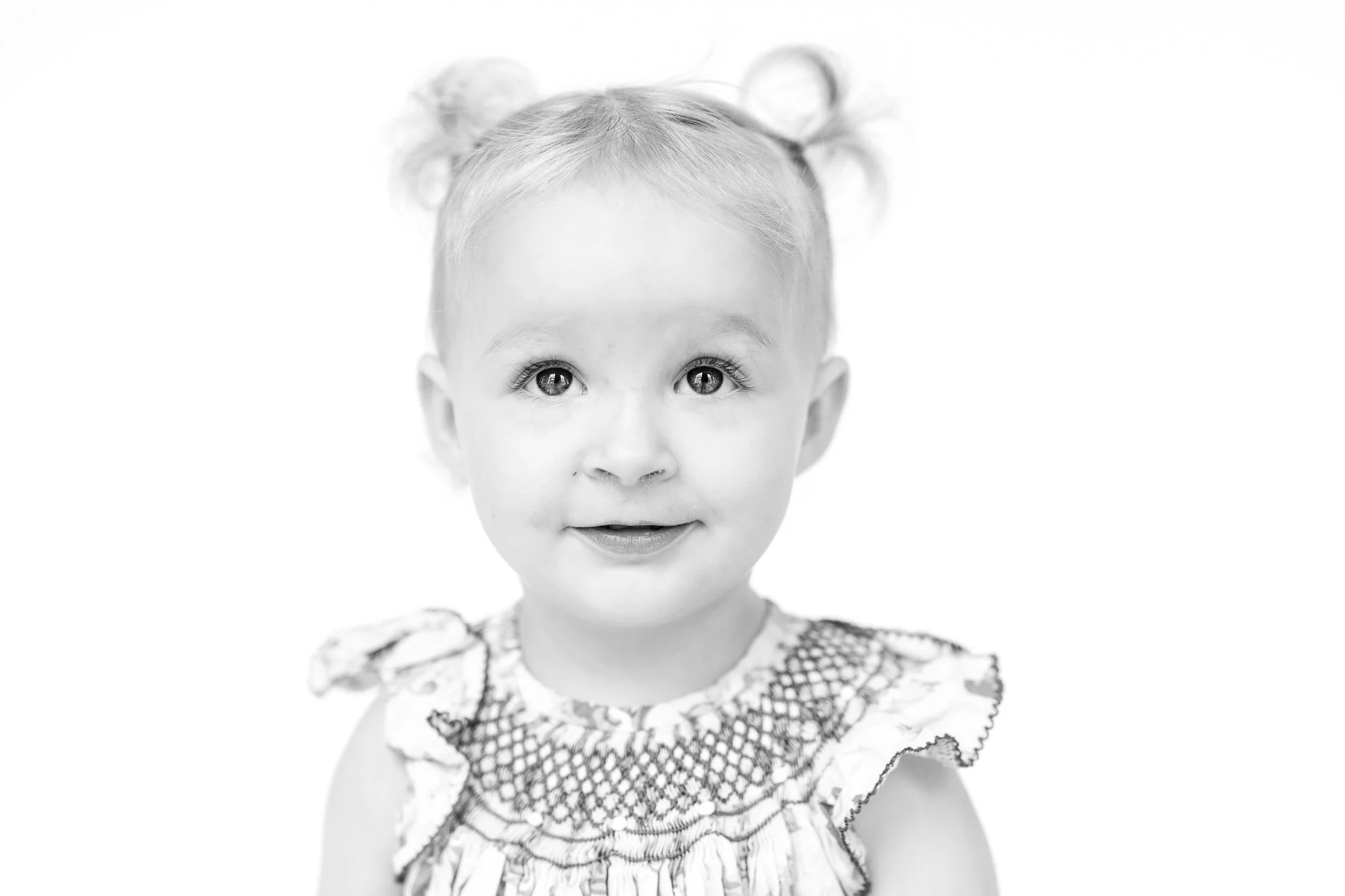 Black and white portrait of a young child with blonde hair in two small buns, wearing a patterned dress.