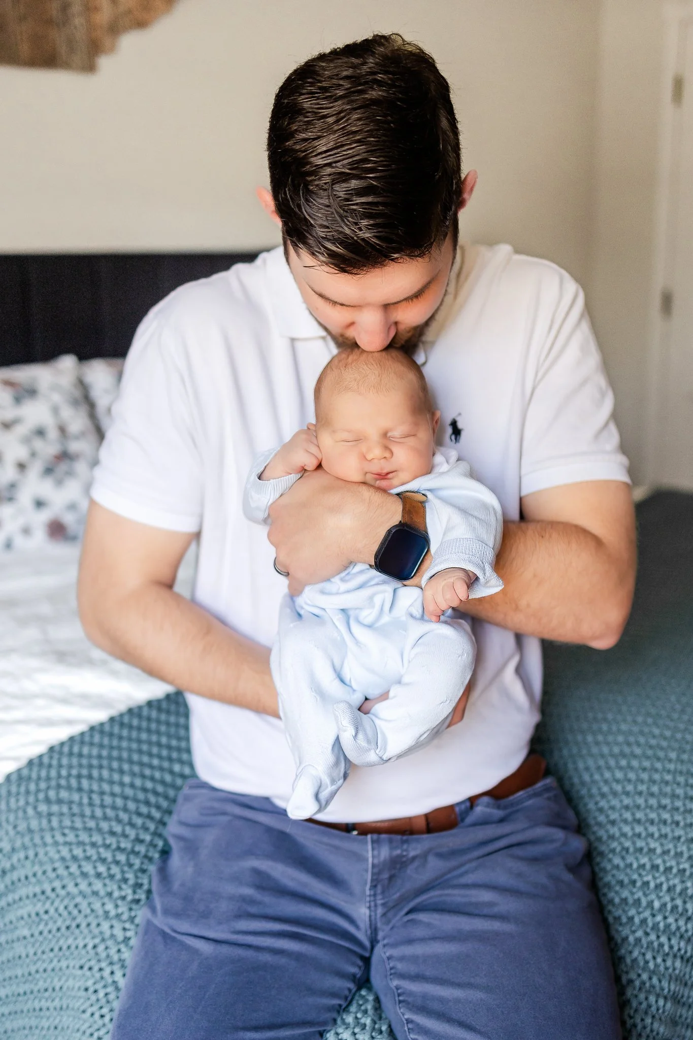 A man gently holding a sleeping baby on his lap, wearing a white shirt and a watch, in a cozy bedroom setting.