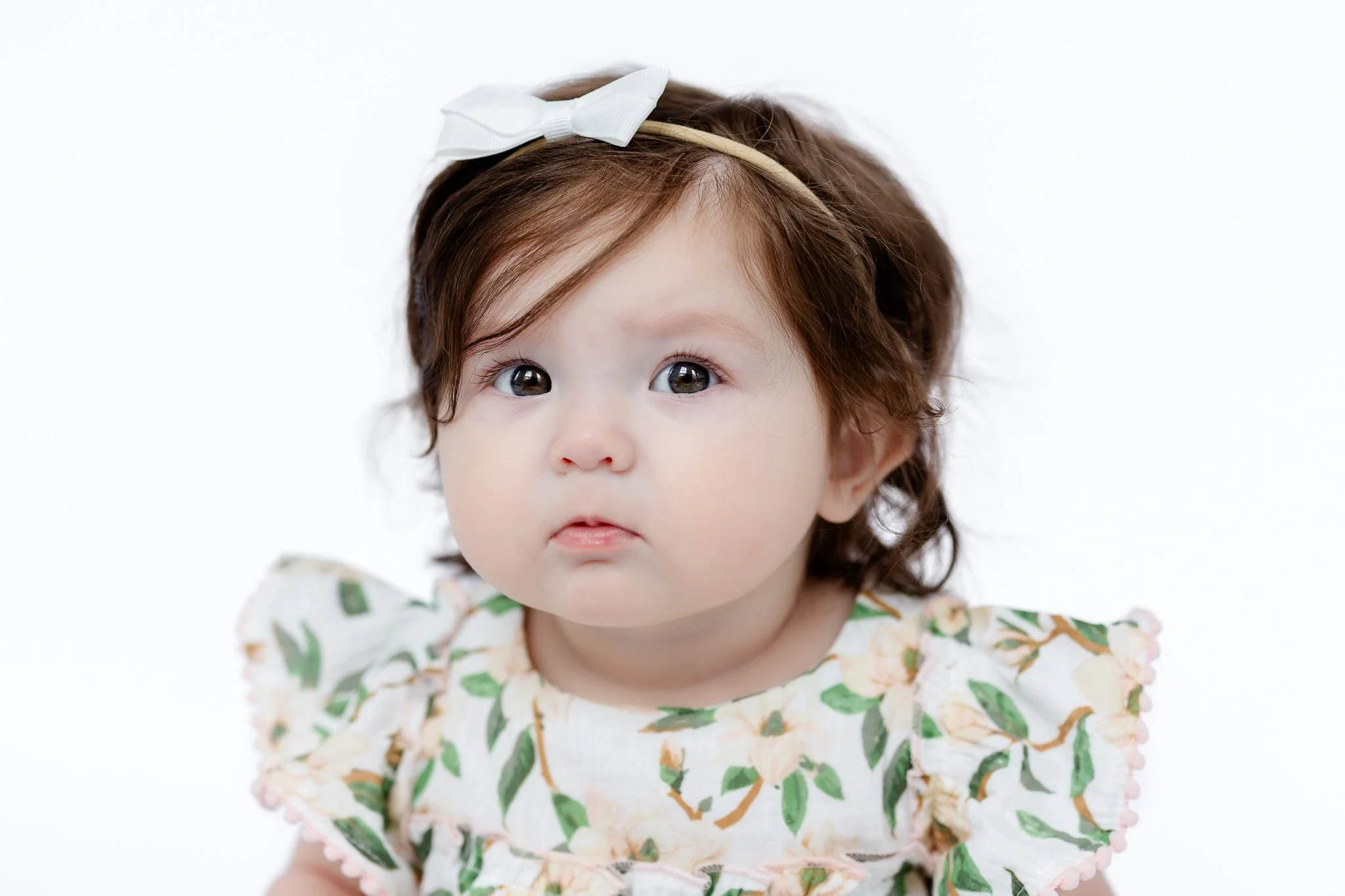 Baby with brown hair and a white bow, wearing a floral dress, looking at the camera against a white background.