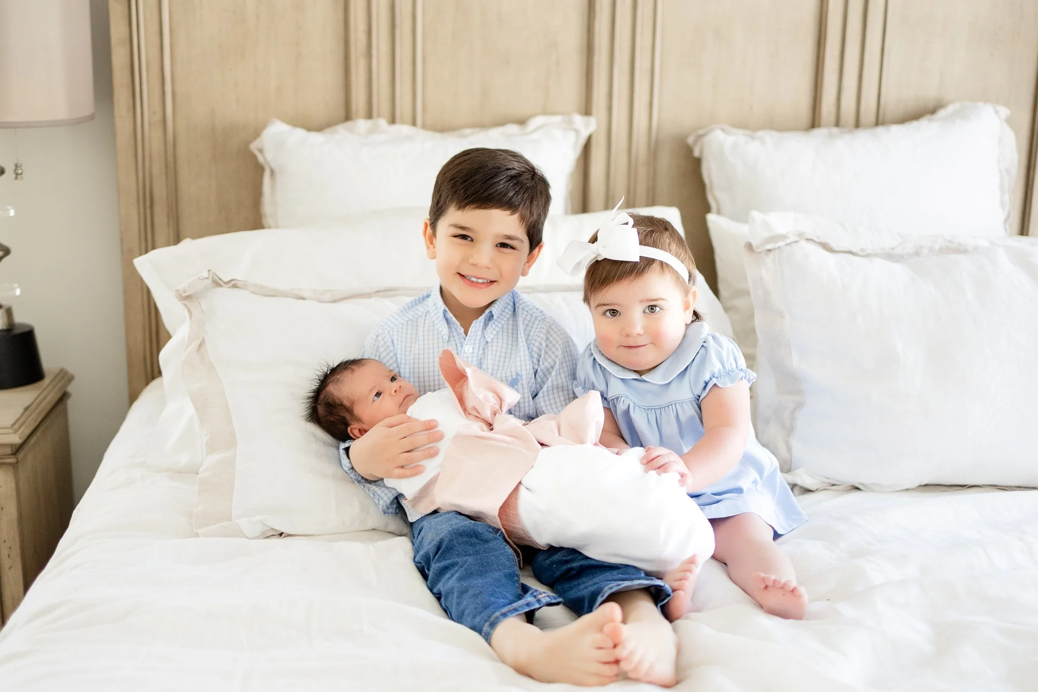 Three children sitting on a bed; an older boy holding a newborn baby wrapped in a blanket, and a toddler girl wearing a blue dress with a white bow.
