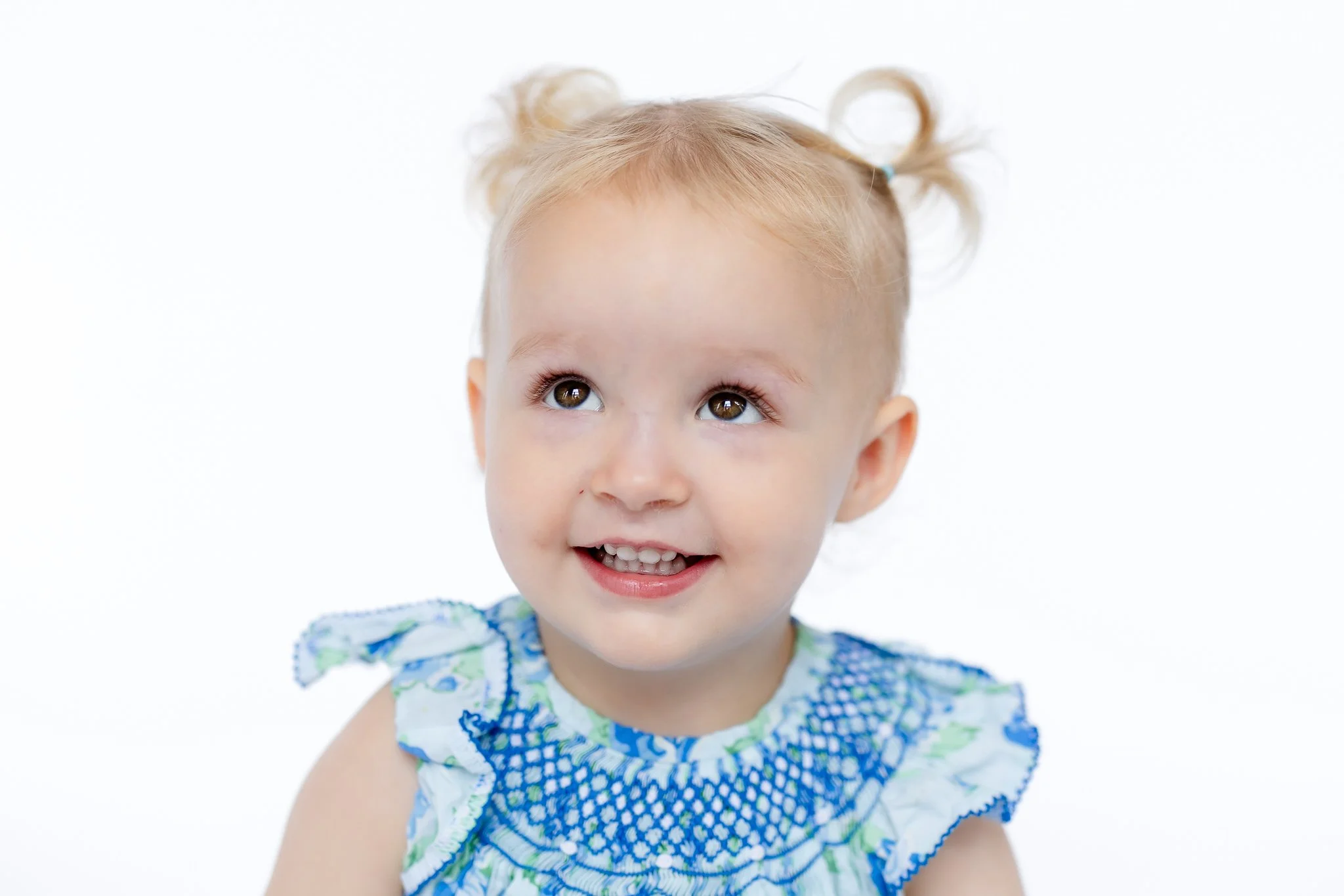A smiling toddler with blonde hair in pigtails, wearing a blue and white patterned dress.
