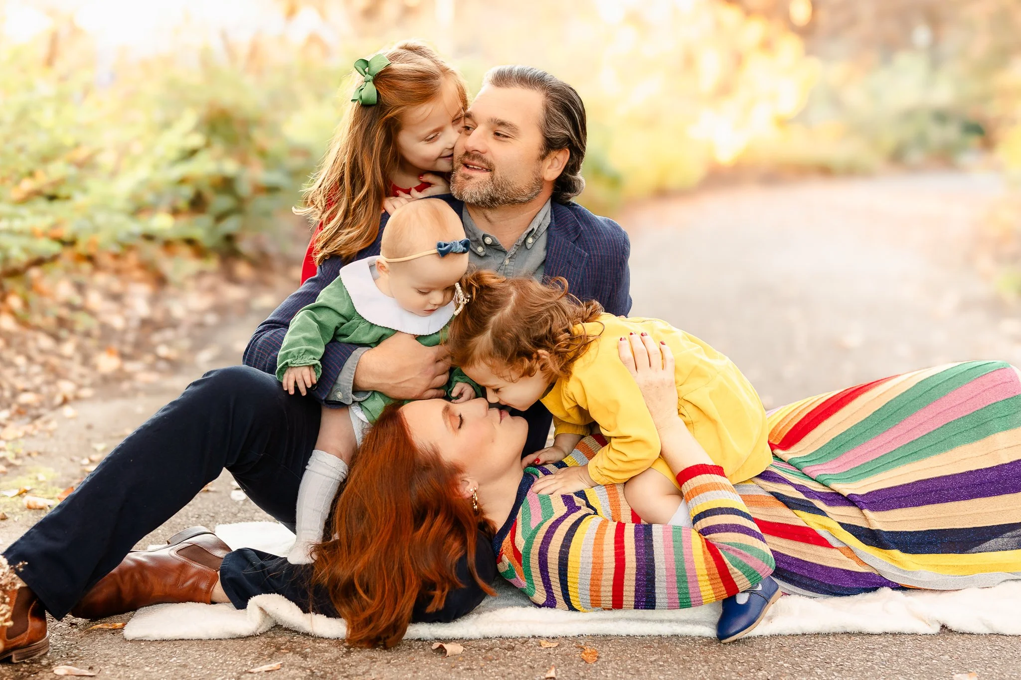 A family with two adults and three children lying on a path surrounded by nature. The mother is lying down in a colorful striped dress with a child in a yellow outfit leaning over her. The father is sitting up, holding a baby in a green outfit. Anoth
