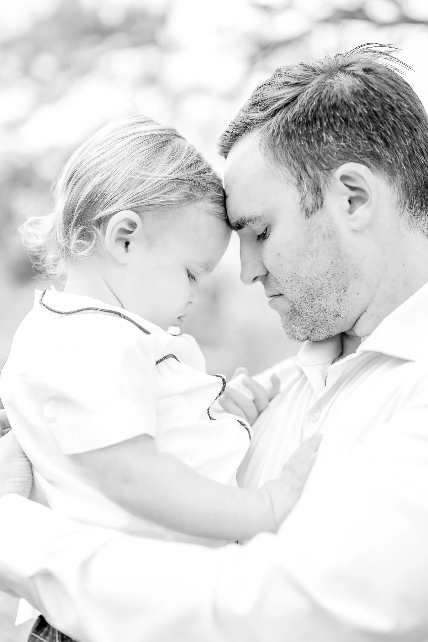 Black and white image of a father gently holding a child, touching foreheads in an intimate pose. The child is wearing a light-colored outfit, and the background is softly blurred.
