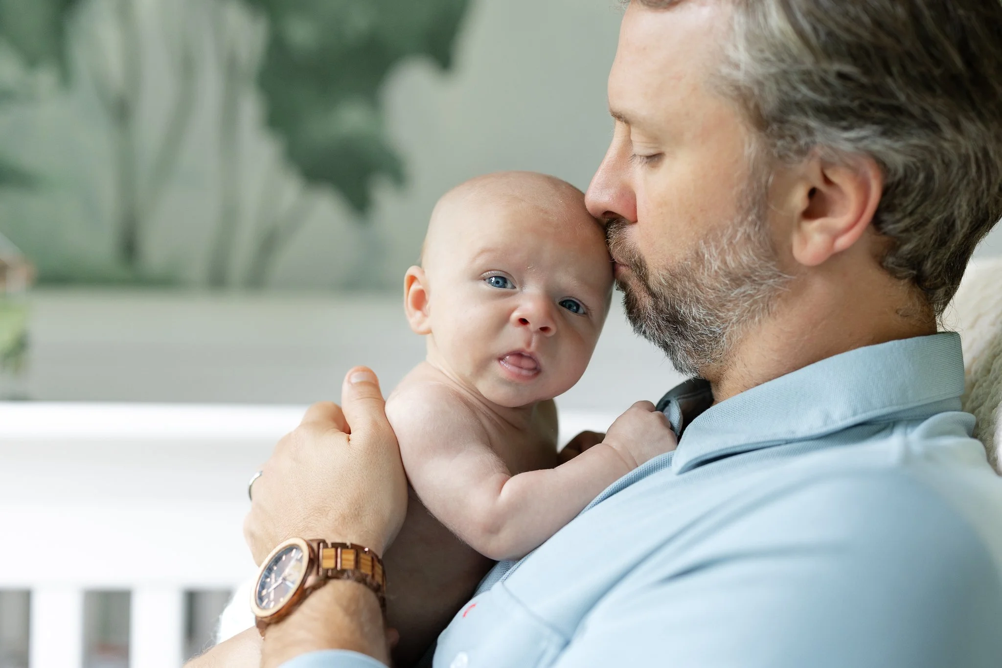 A bearded man in a light blue shirt holding and kissing a baby, indoors.