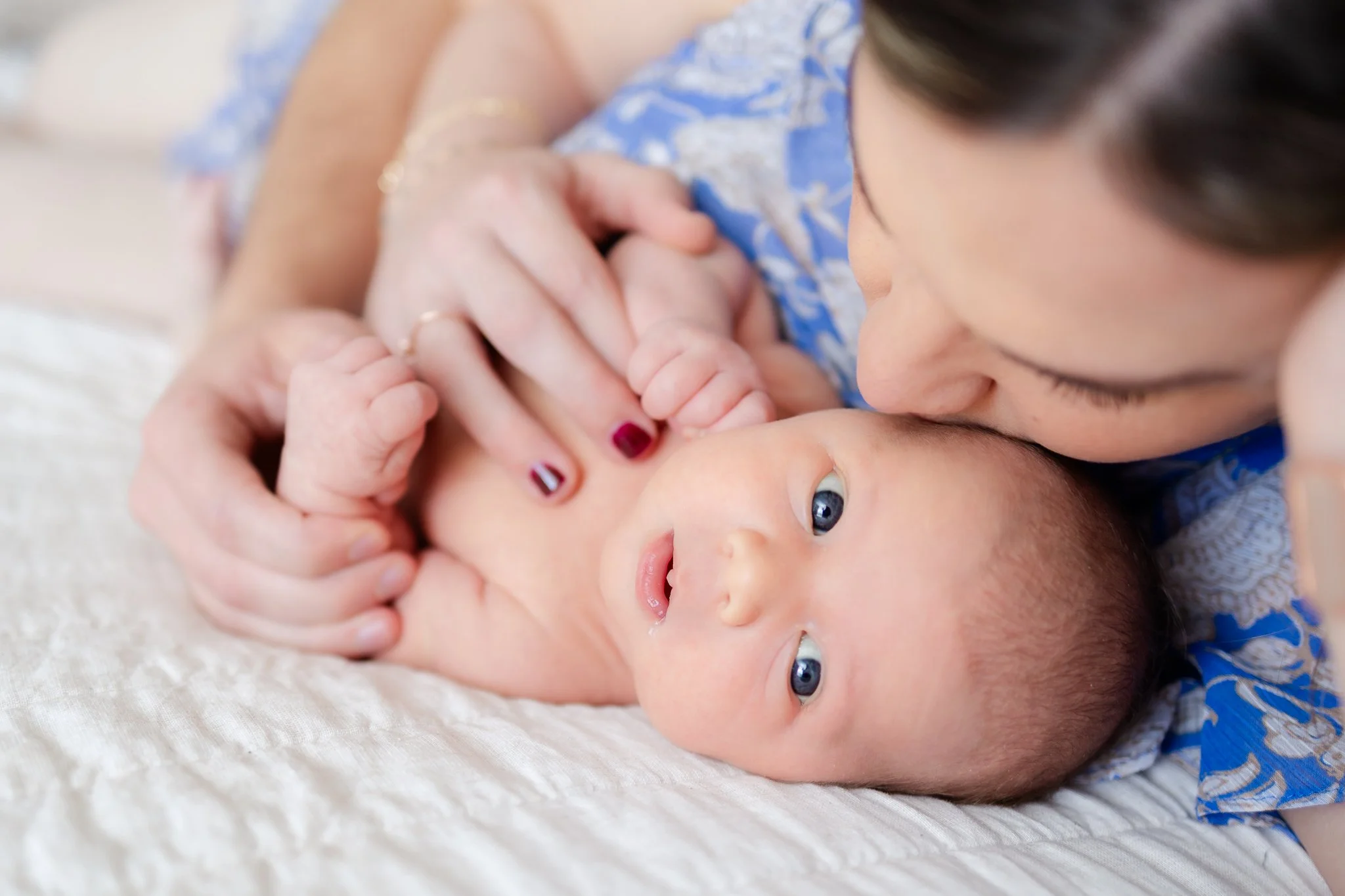 Mother gently kissing her baby's forehead while holding the baby's hand on a white blanket.