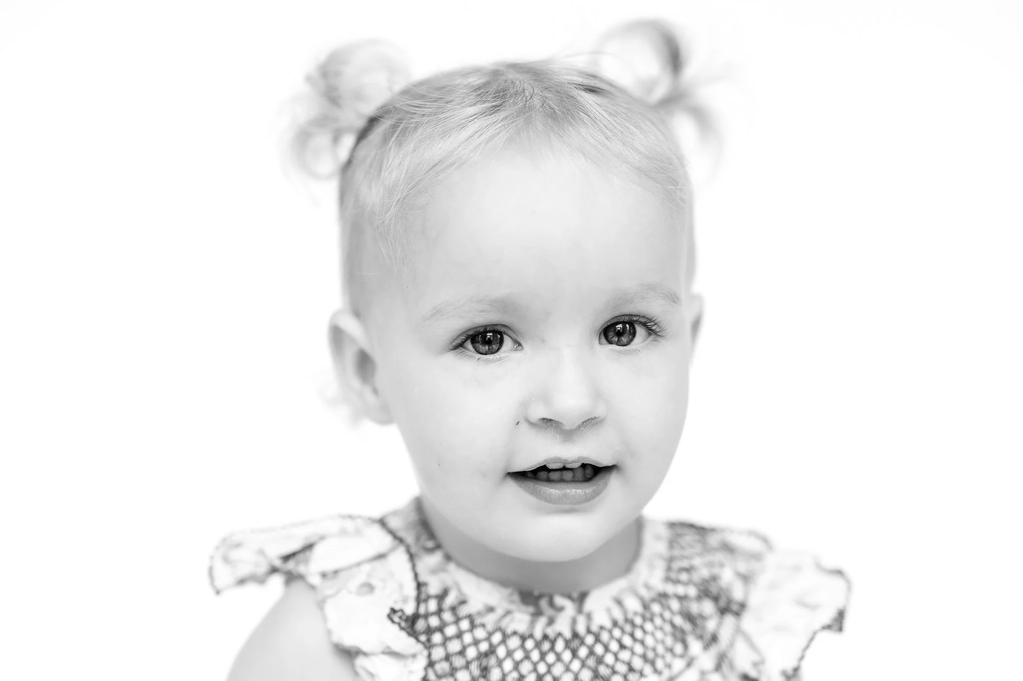 Black and white portrait of a toddler with light hair in pigtails, wearing a patterned top, smiling softly against a plain background.