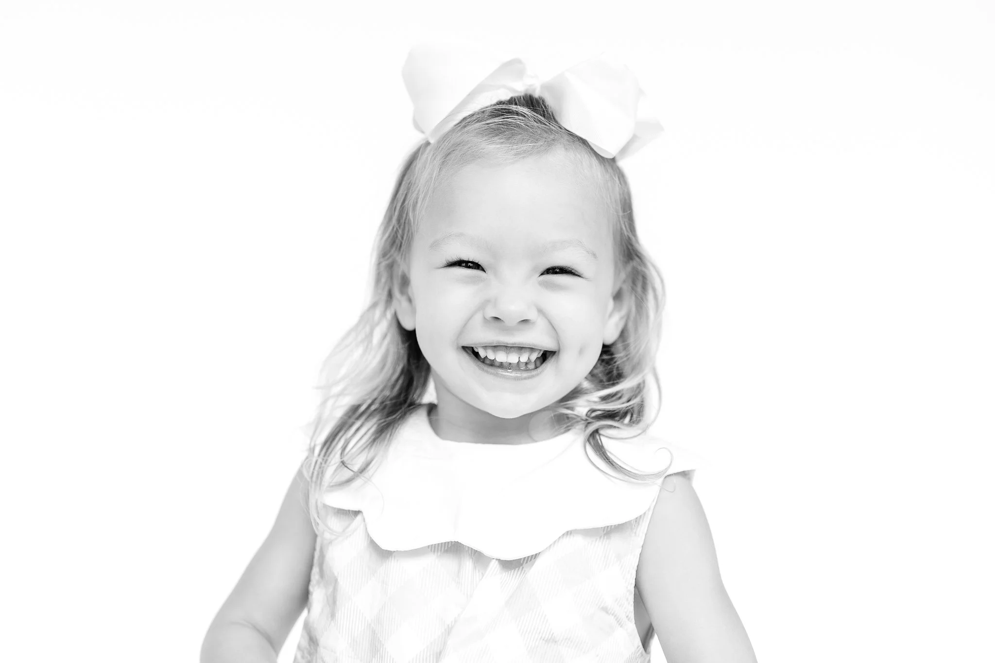 Black and white photo of a smiling toddler with a large bow in her hair, wearing a sleeveless dress.