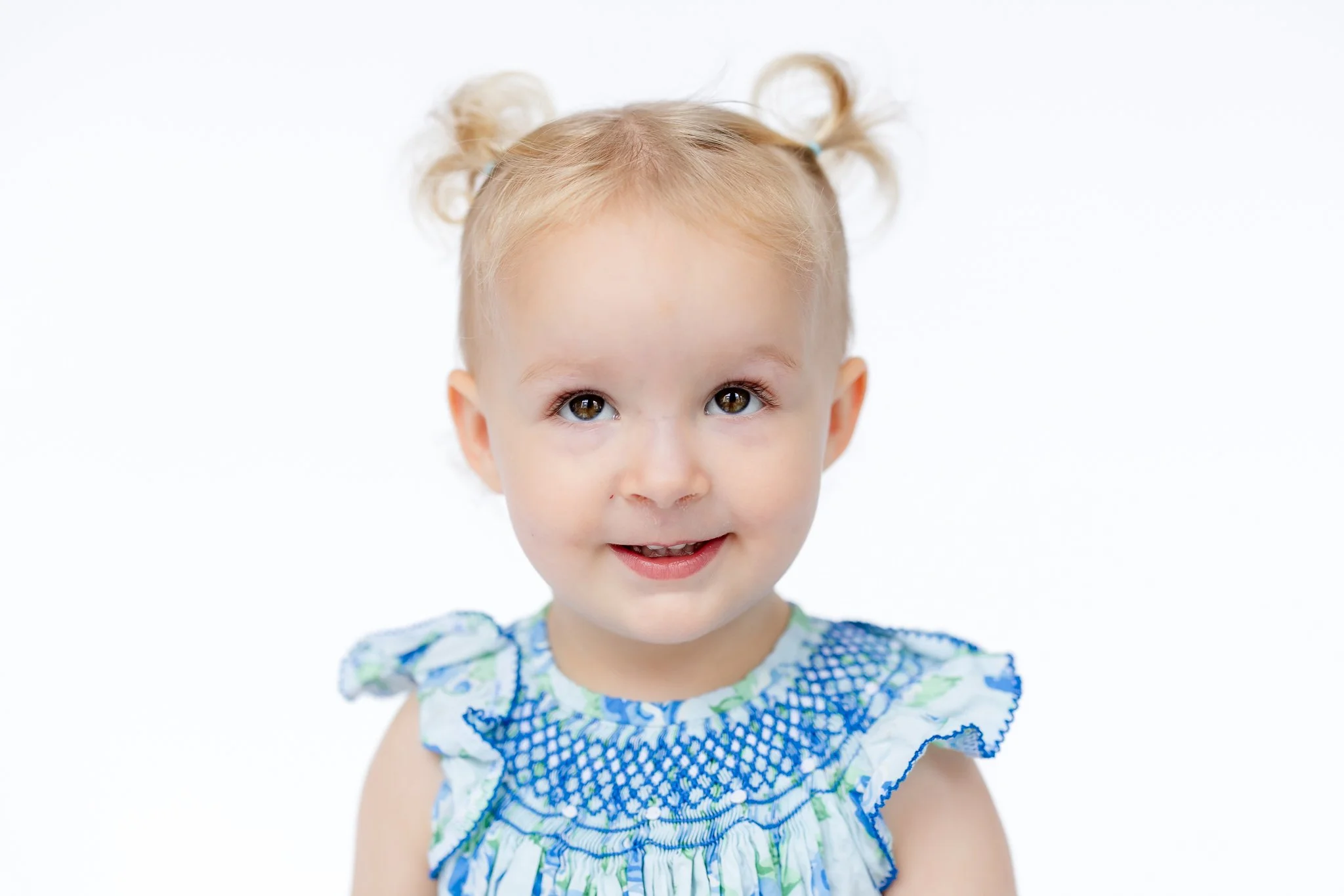 Smiling toddler with two small pigtails wearing a blue patterned dress on a white background.