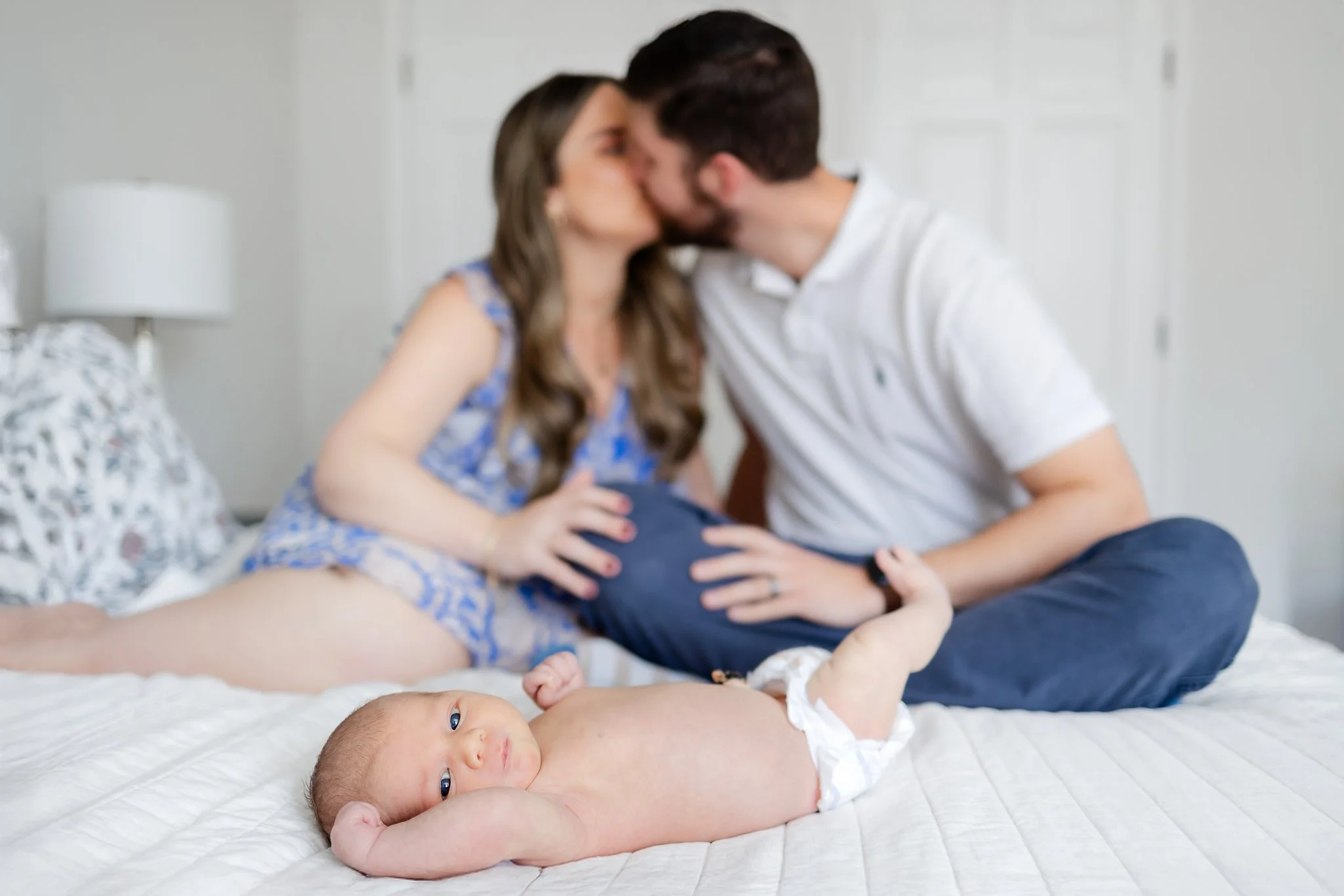 Baby lying on bed in foreground, couple kissing in background.