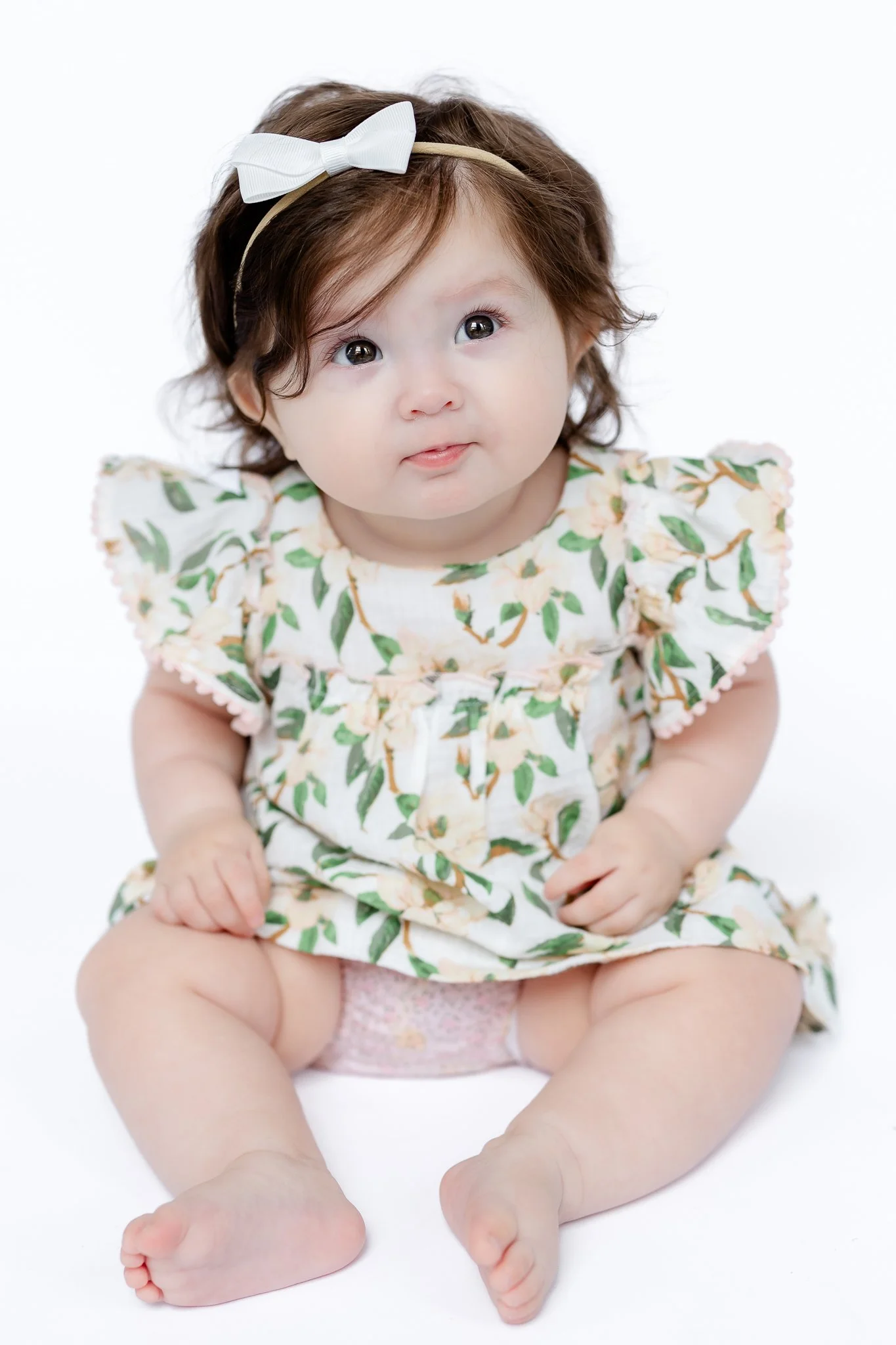 Baby wearing floral dress and white bow headband on light background.