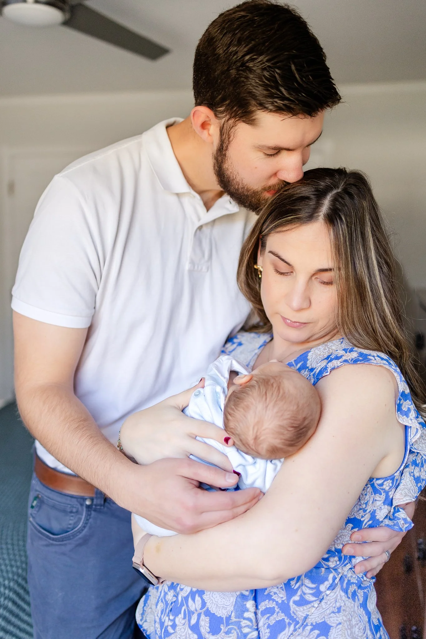 A couple lovingly holding their newborn baby indoors, with the father kissing the mother on the head.