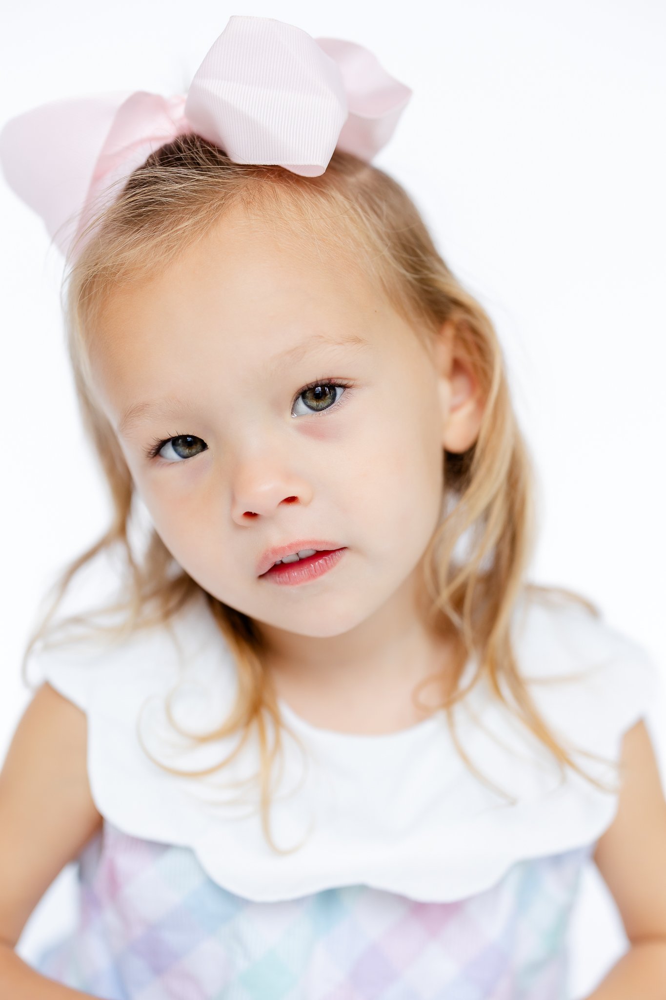 Young girl wearing pink bow and pastel dress, looking softly at camera.