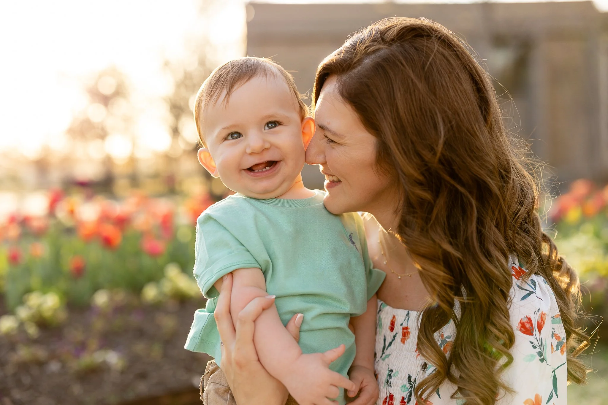 A mother holding her smiling baby in a garden with flowers in the background.
