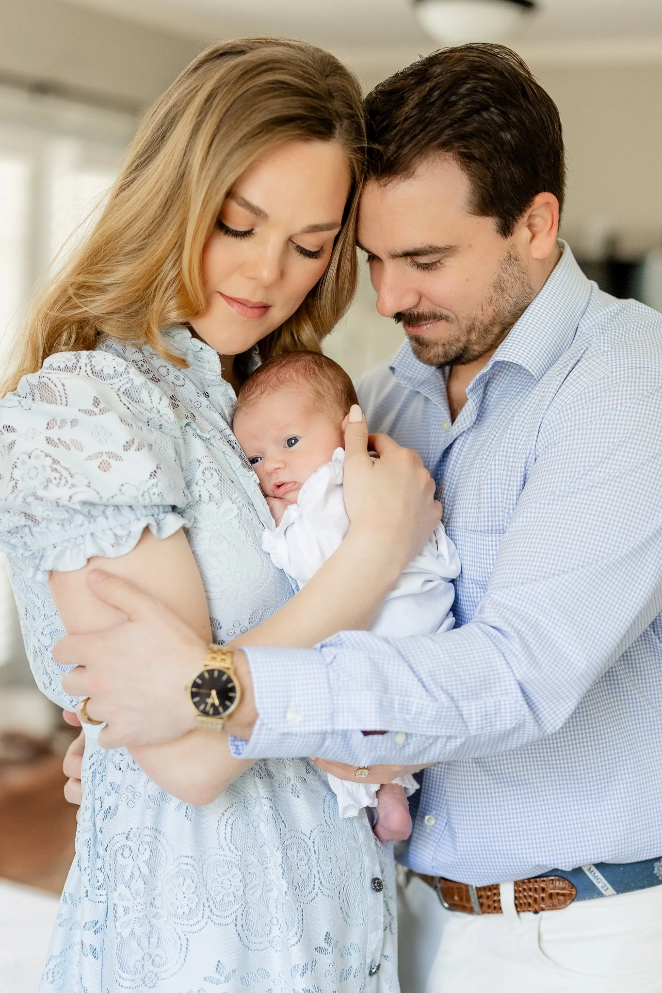 A couple lovingly holds their newborn baby in a bright room, with both parents wearing light-colored clothing. The mother is in a lace dress, and the father in a checkered shirt.